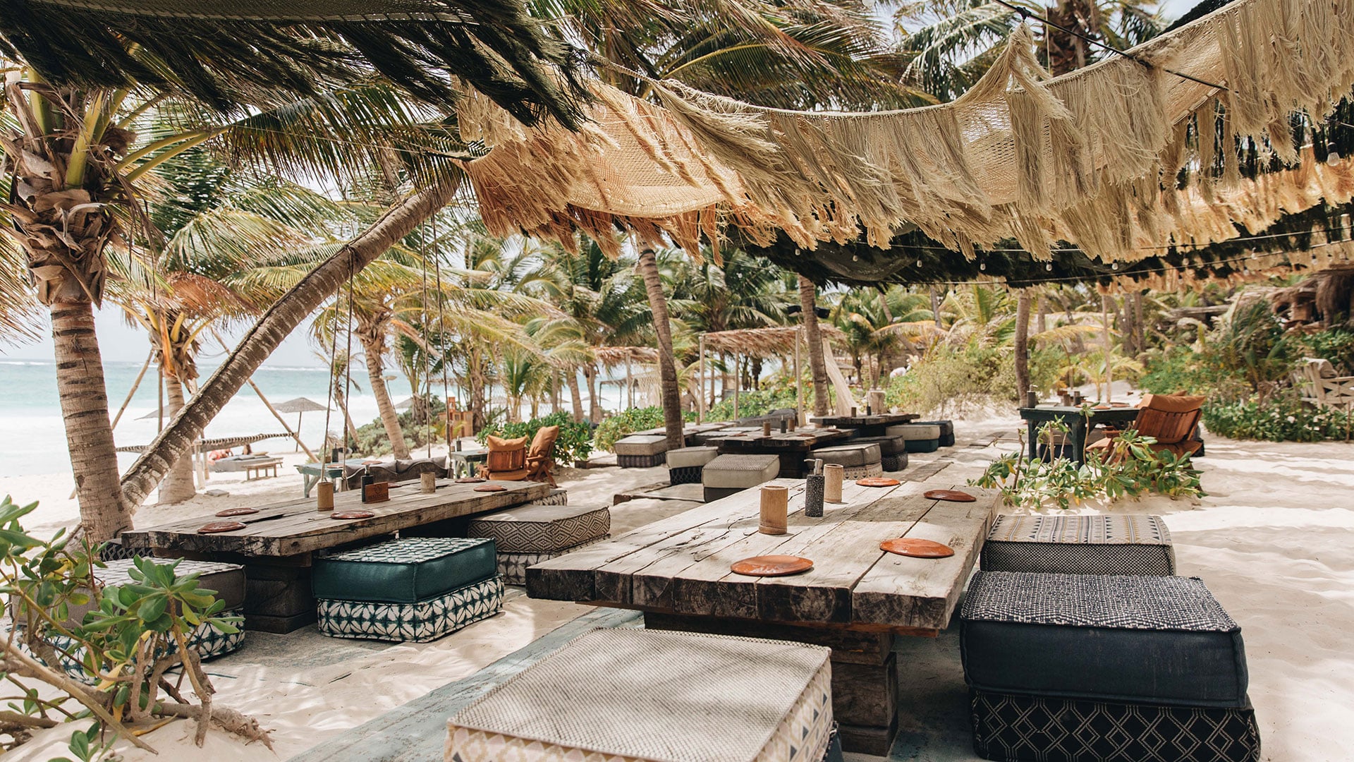 La Popular, Mexico — Beachfront dining pavilion with woven palm shade structures, wooden tables, black cushioned seating, and turquoise ocean views beneath coconut palms.