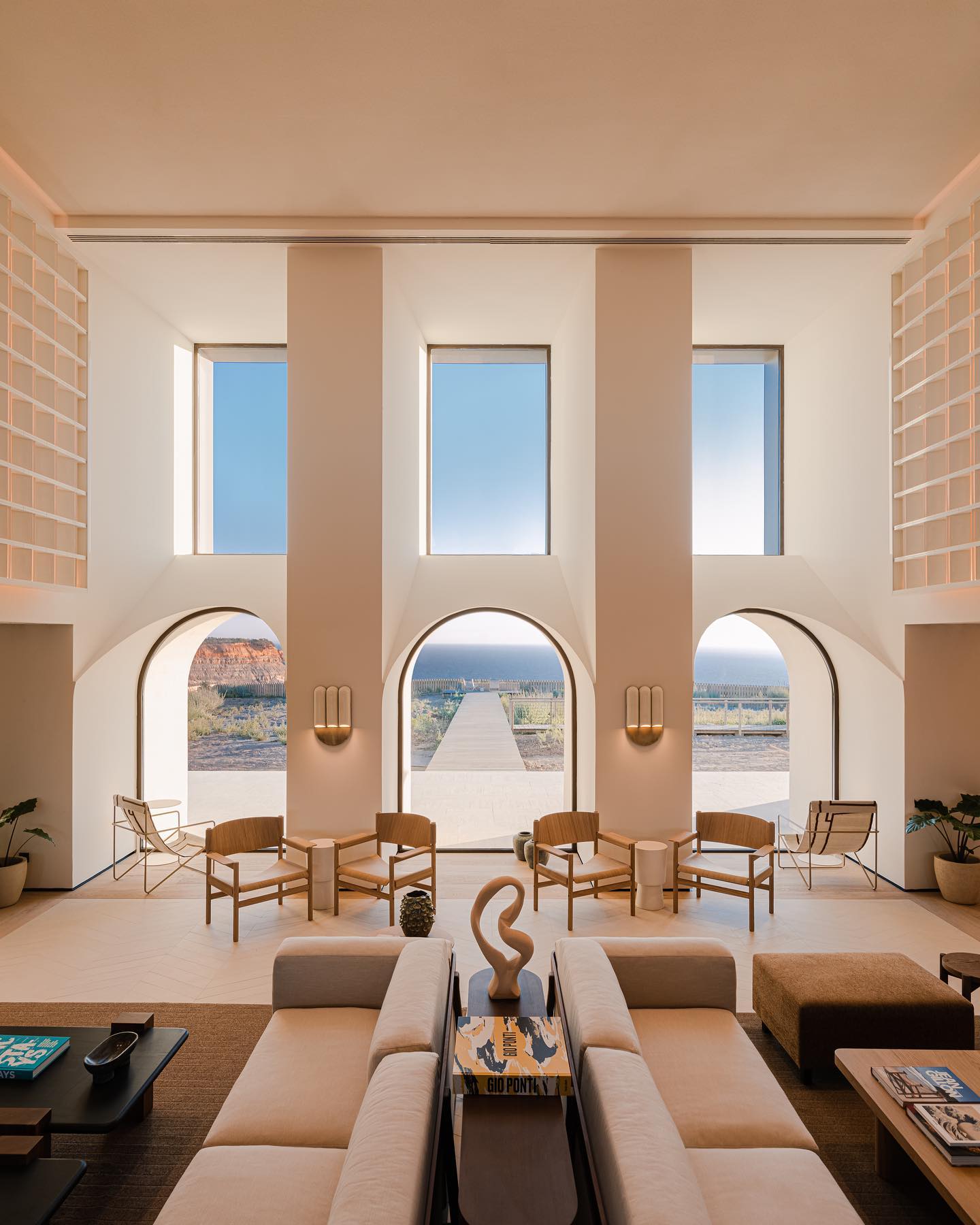 Aethos Ericeira, Portugal — Minimalist lounge with arched doorways, warm earth tones, wooden chairs, and panoramic views of coastal landscape through floor-to-ceiling windows.