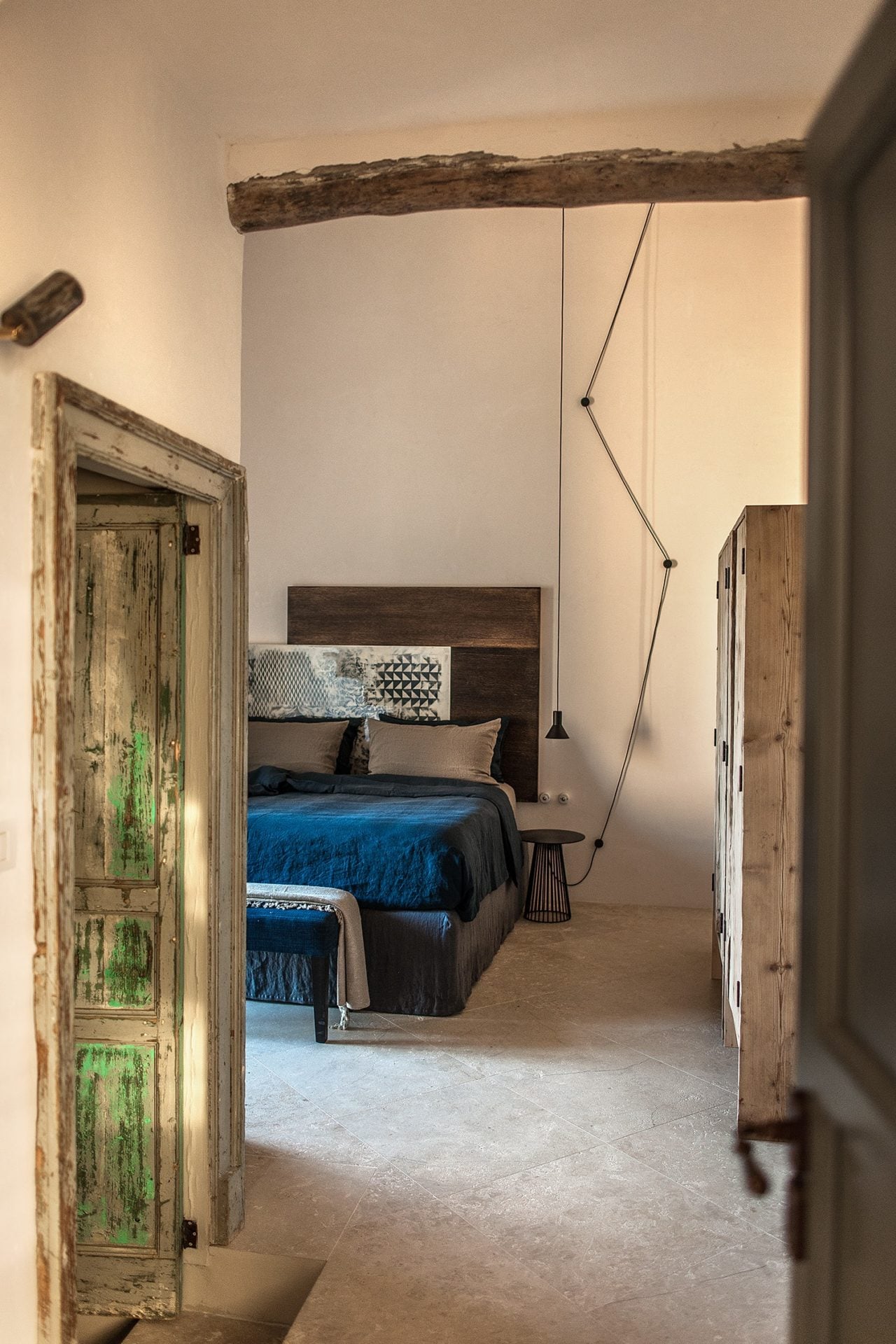 Dimora delle Balze, Europe — Rustic wooden doors frame minimalist bedroom with exposed beam, blue bedding, pendant lighting, and weathered terracotta floors.