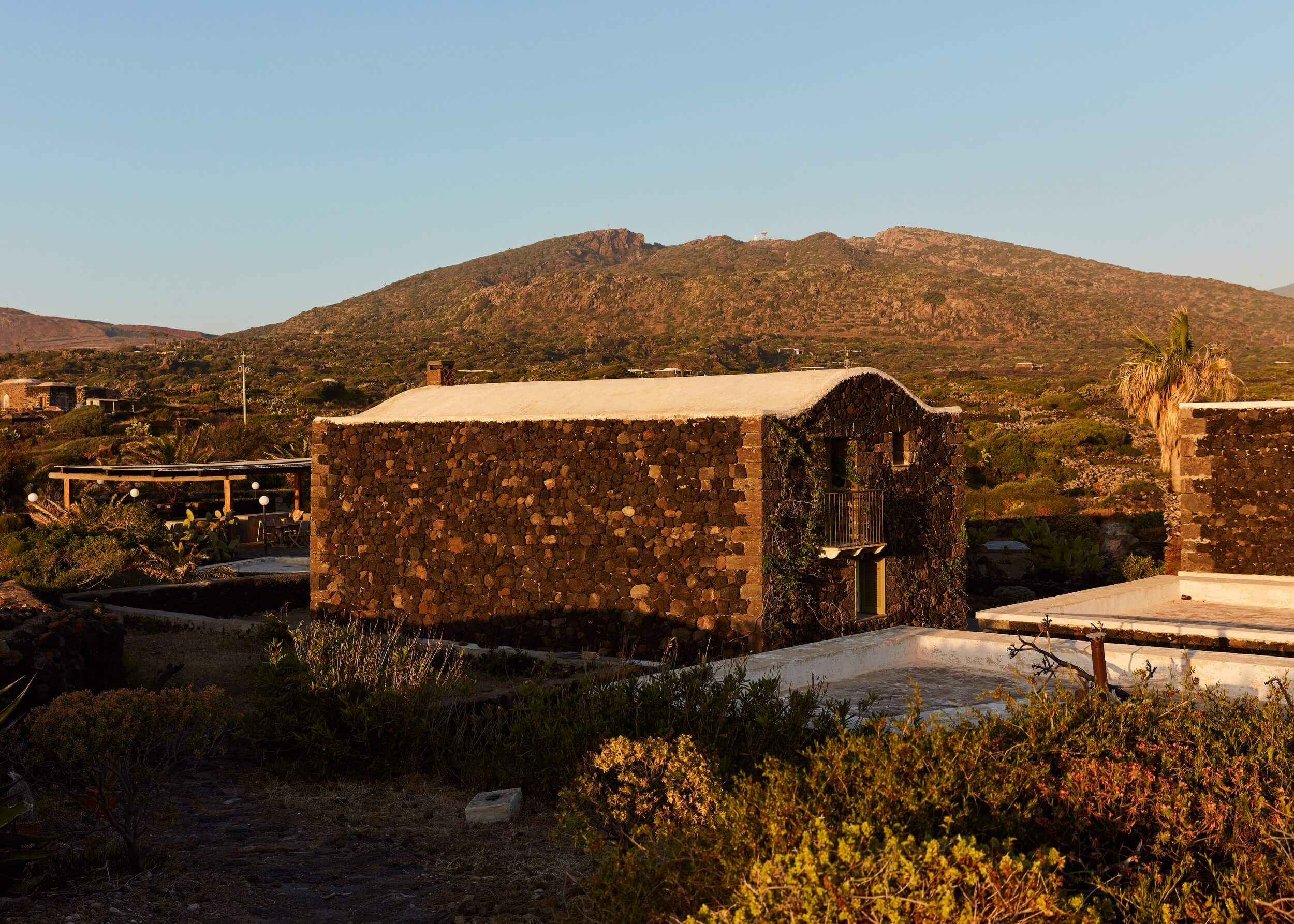 Parco Dei Sesi, Europe — Stone dwelling with arched roof beneath dramatic hillside, desert landscape, golden hour light casting warm shadows across volcanic rock architecture.