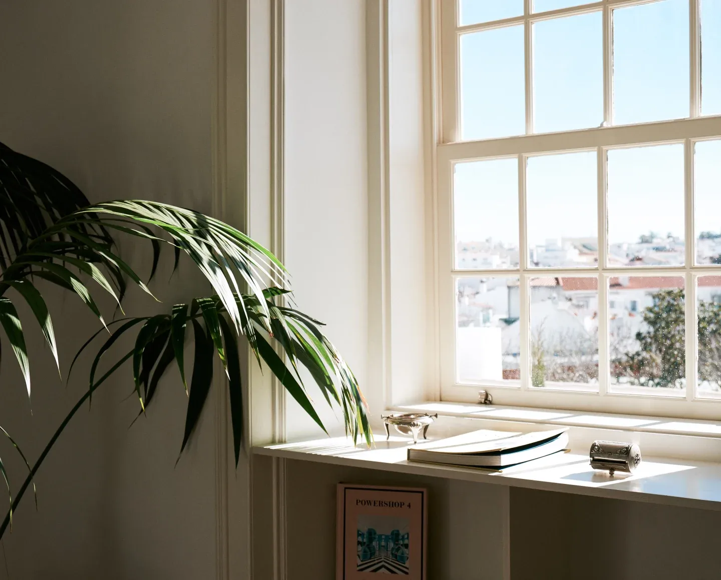 Casa Mãe, Europe — Bright corner office with large gridded windows, green potted palm, open notebooks, and cityscape view in soft natural light.