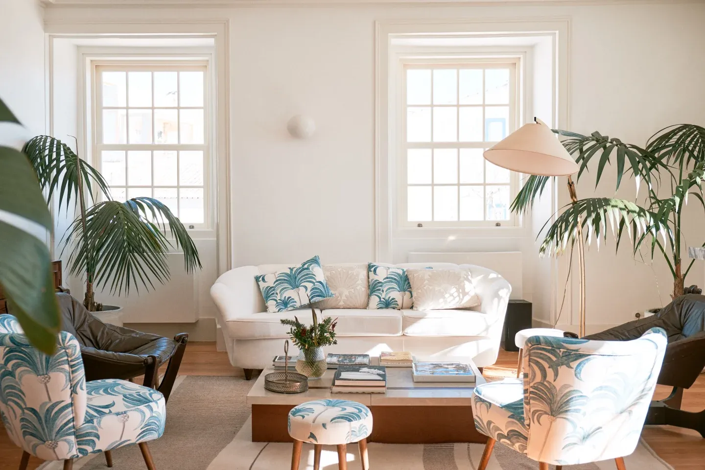 Casa Mãe, Europe — Sunlit living room with curved white sofa, palm plants, blue patterned chairs, wooden coffee table, and grid-pane windows.