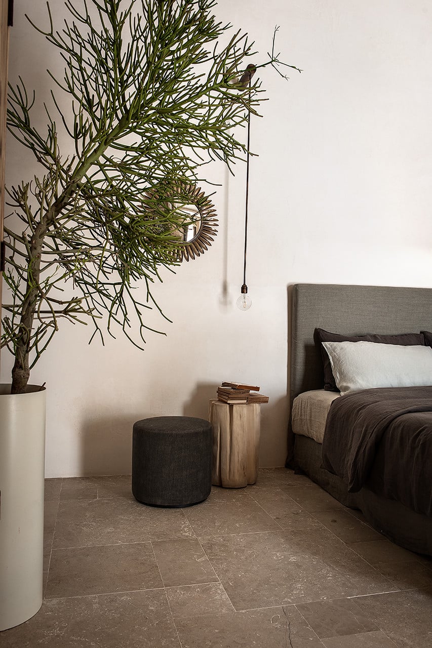 Dimora delle Balze, Europe — Minimalist bedroom with concrete flooring, potted tree, dark upholstered bed, wooden side table, charcoal pouf, and pendant light.