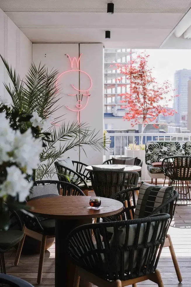 Loreta, Europe — Modern rooftop terrace with black woven chairs, wooden table, pink neon signage, potted plants, and cityscape views.