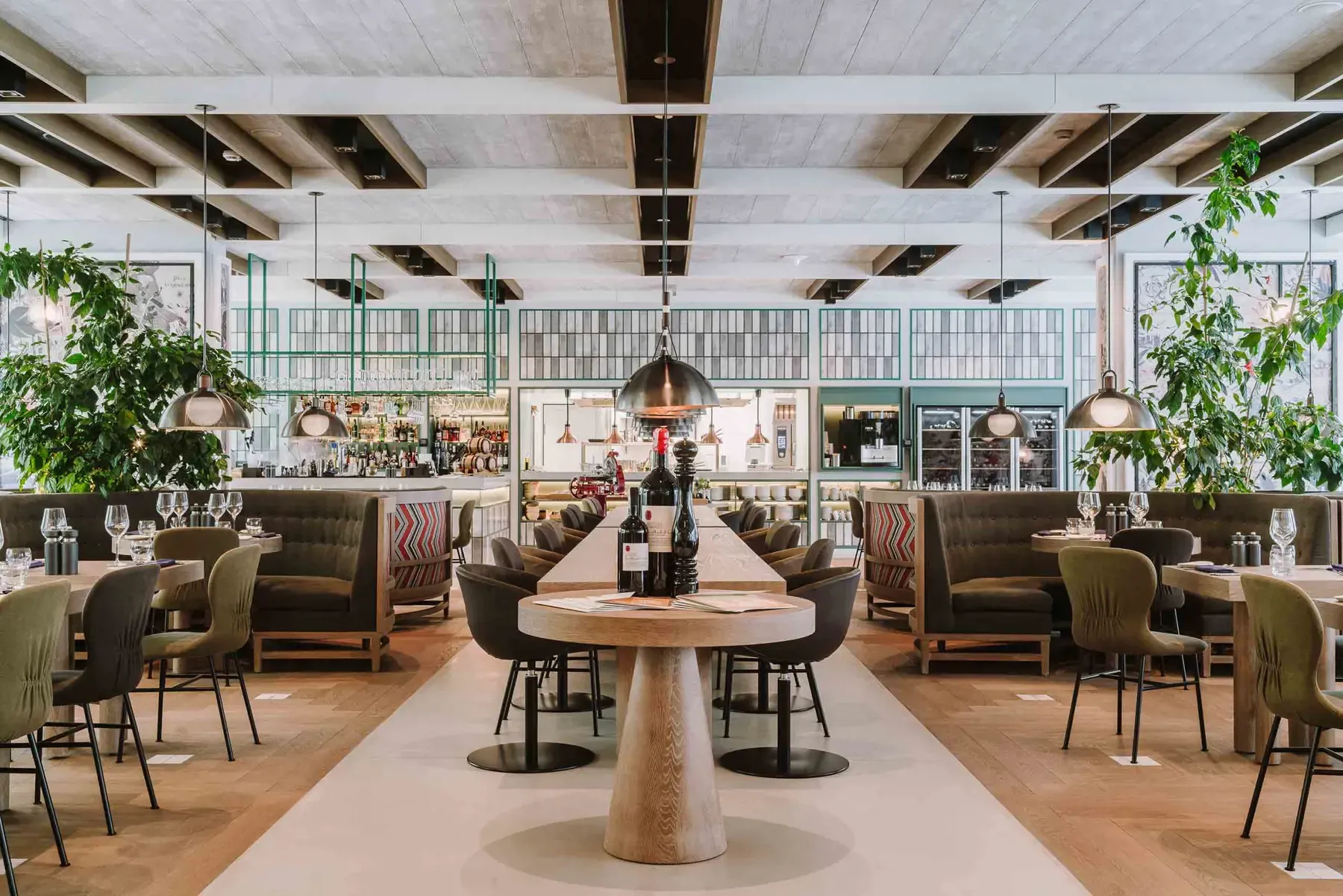 Magari, Europe — Modern restaurant with coffered ceiling, pendant lights, wooden pedestal tables, olive leather banquettes, and verdant hanging plants creating sophisticated hospitality design.