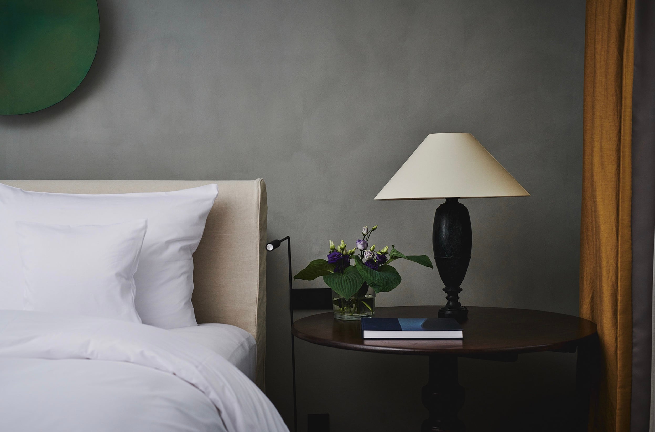 Purs, Europe — Modern luxury bedroom nightstand with black lamp, purple flowers, dark wood table, cream upholstered headboard, gray wall, warm lighting.