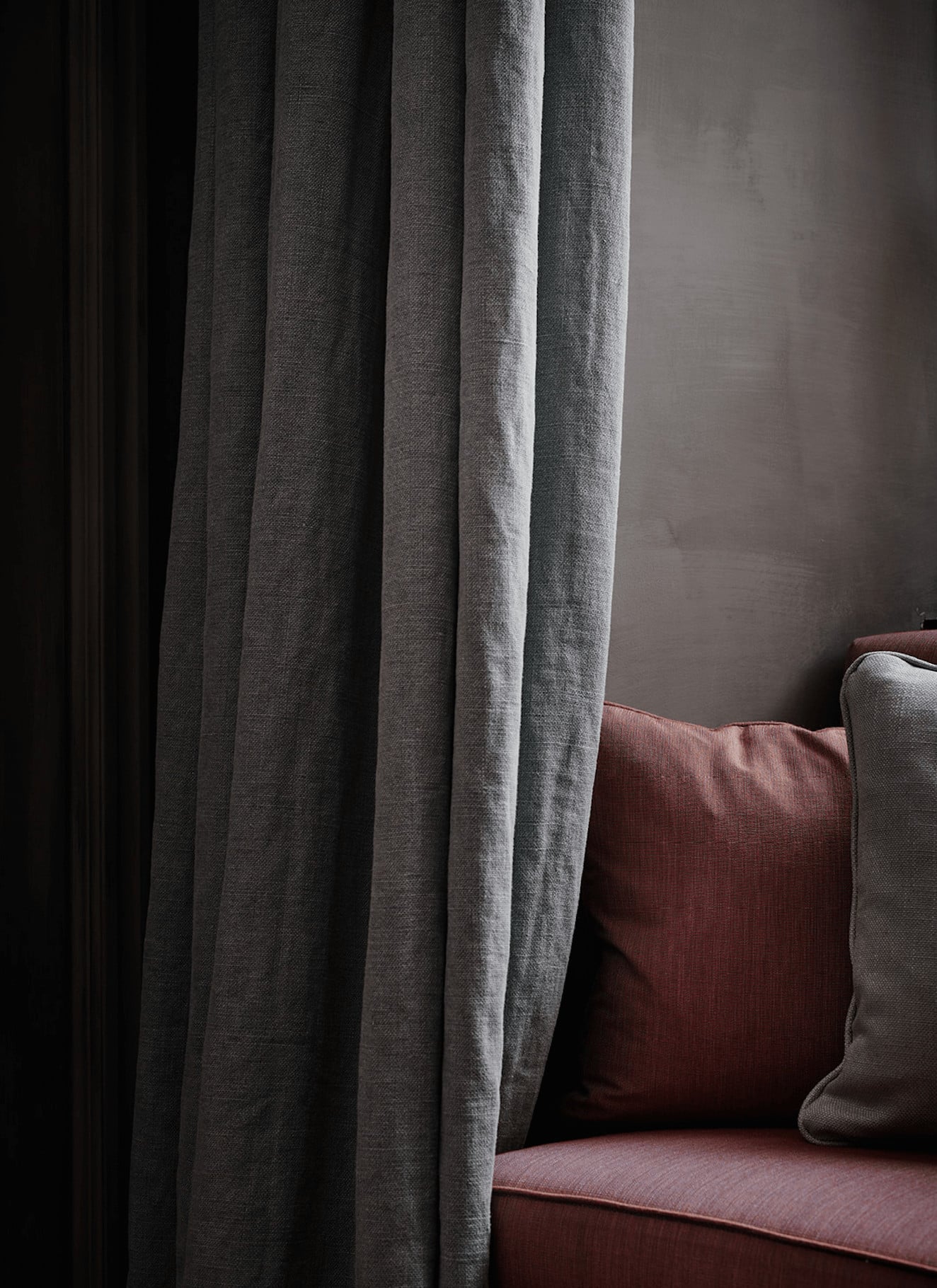 Purs, Europe — Charcoal linen curtains and burgundy upholstered sofa with cream pillow in minimalist interior with soft natural light.