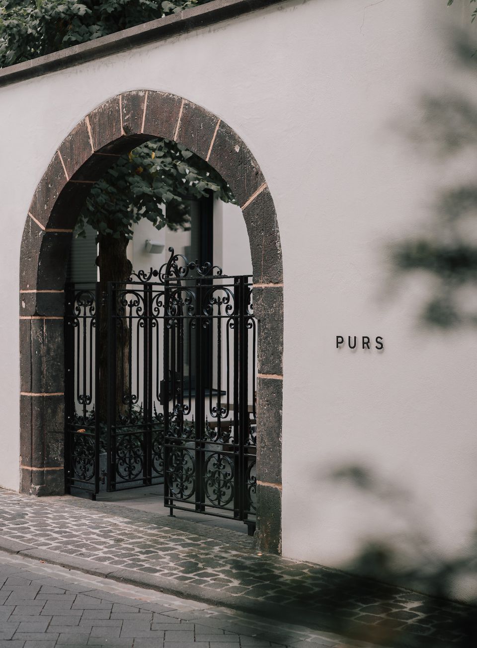 Purs, Europe — Arched stone doorway with ornate black ironwork gate and balcony, white stucco facade, dappled shadows, minimalist luxury architecture.