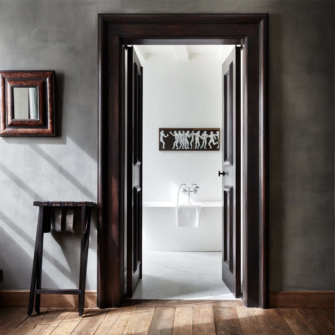 Purs, Europe — Dark wood double doors open to minimalist bathroom with classical frieze relief, marble floors, and sculptural bath fixtures in natural light.