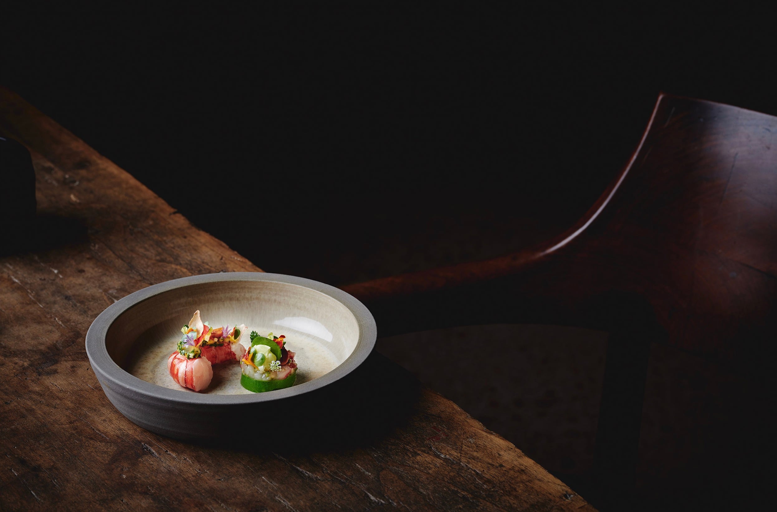 Purs, Europe — Minimalist ceramic bowl with delicate plated dish on dark wood surface, architectural chair silhouette, dramatic black backdrop, refined fine dining aesthetic.