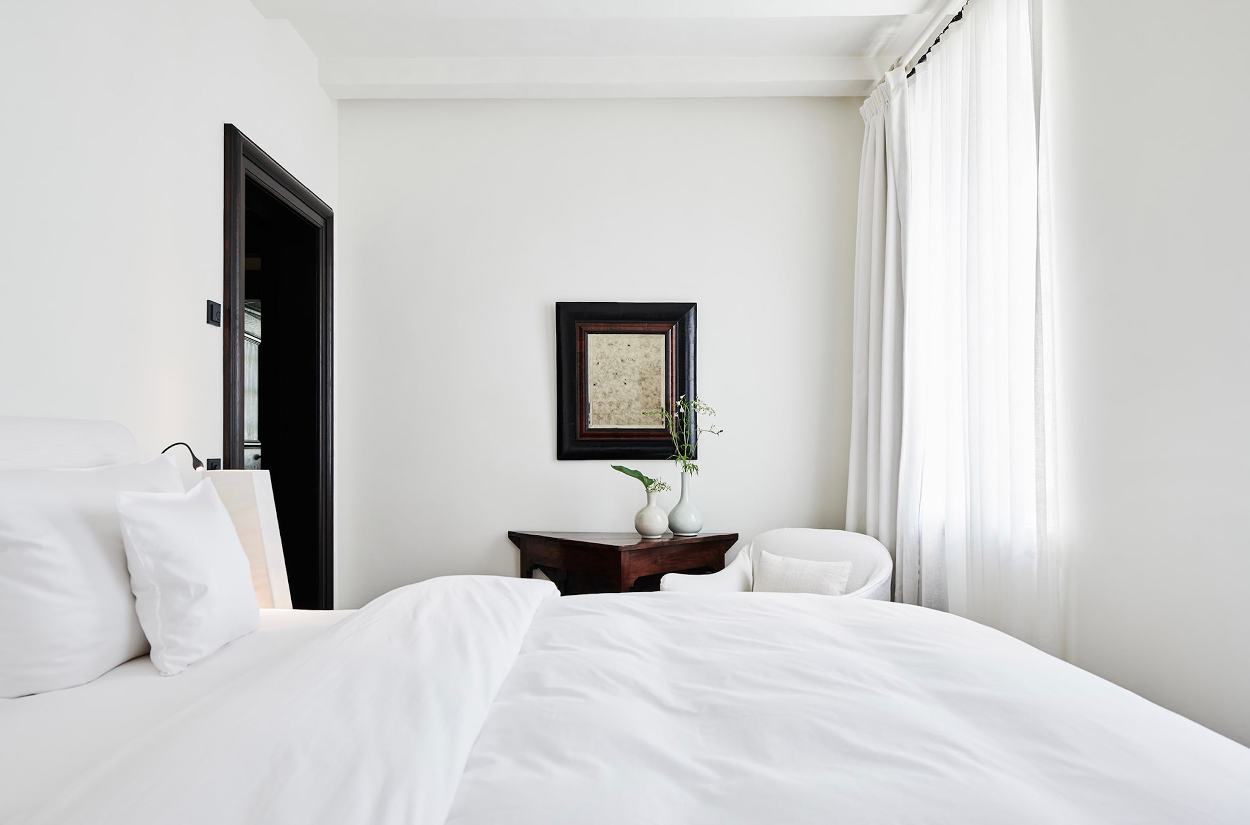 Purs, Europe — Minimalist bedroom with white linens, dark wood nightstand, framed mirror, potted flowers, and sheer curtains filtering natural light.