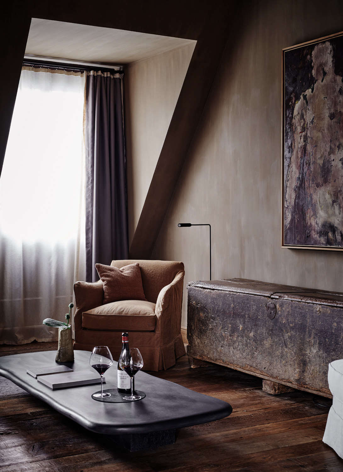 Purs, Europe — Minimalist suite with angled ceiling, linen armchair, dark wood flooring, framed abstract artwork, floor lamp, wine service, and dramatic natural light from tall window.