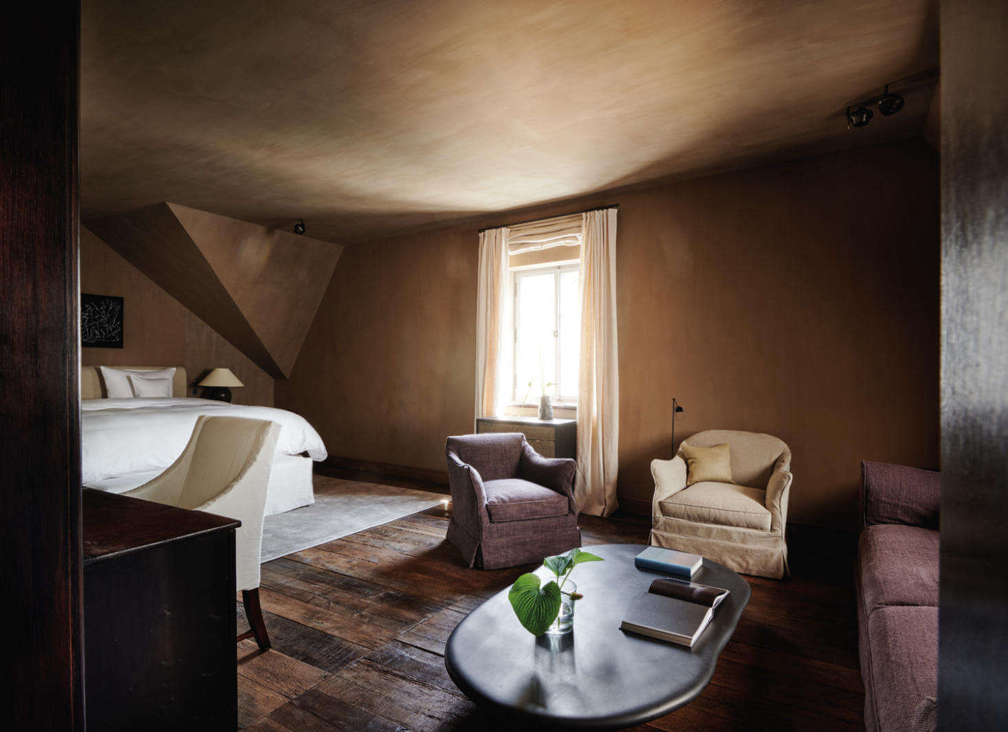 Purs, Europe — Warm earth-toned bedroom suite with angled ceiling, upholstered seating, hardwood floors, natural light, and minimalist modern furnishings.