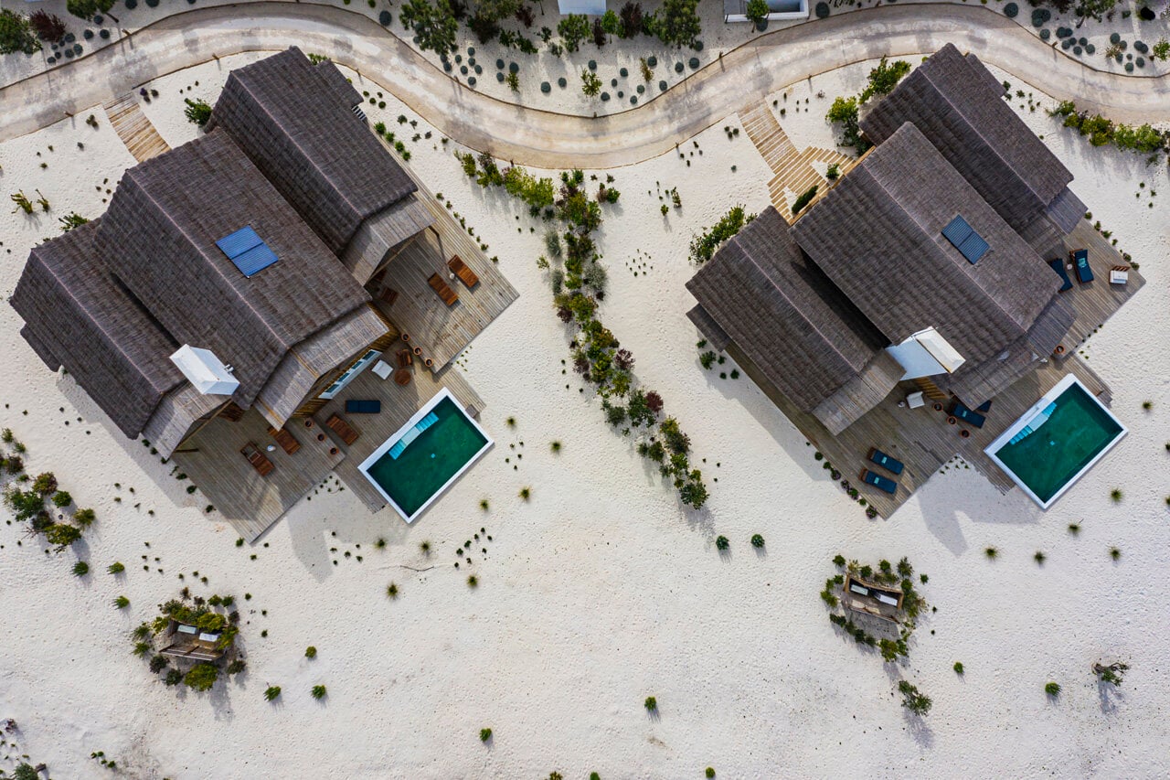 Quinta da Comporta, Europe — Aerial view of two modernist villas with dark roofs, wooden decks, turquoise pools, and sculptural landscaping on white sand.