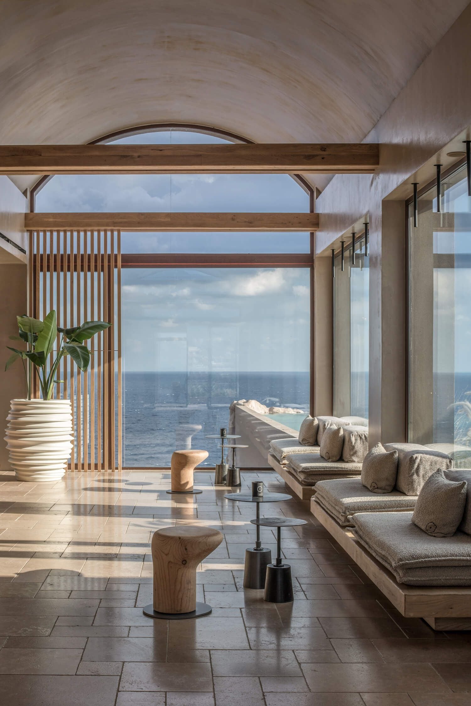 ACRO Suites, Europe — Minimalist coastal lounge with barrel-vault ceiling, floor-to-ceiling ocean views, timber beams, linen seating, and natural light casting geometric shadows on travertine floors.