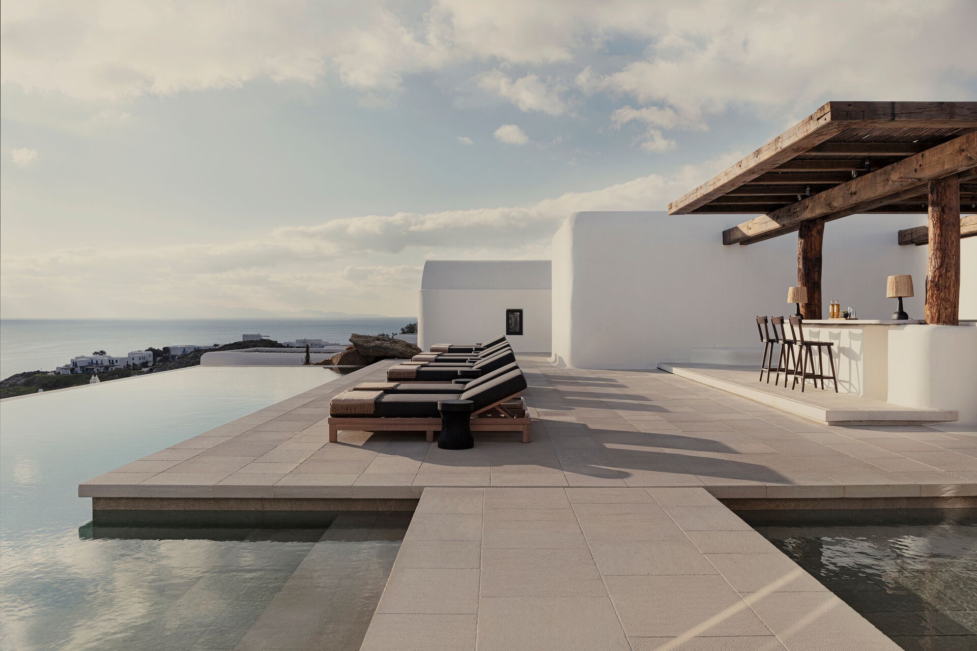 Kalesma, Europe — Minimalist clifftop terrace with infinity pool, striped loungers, wooden pergola bar, and calm sea horizon beneath clouded sky.