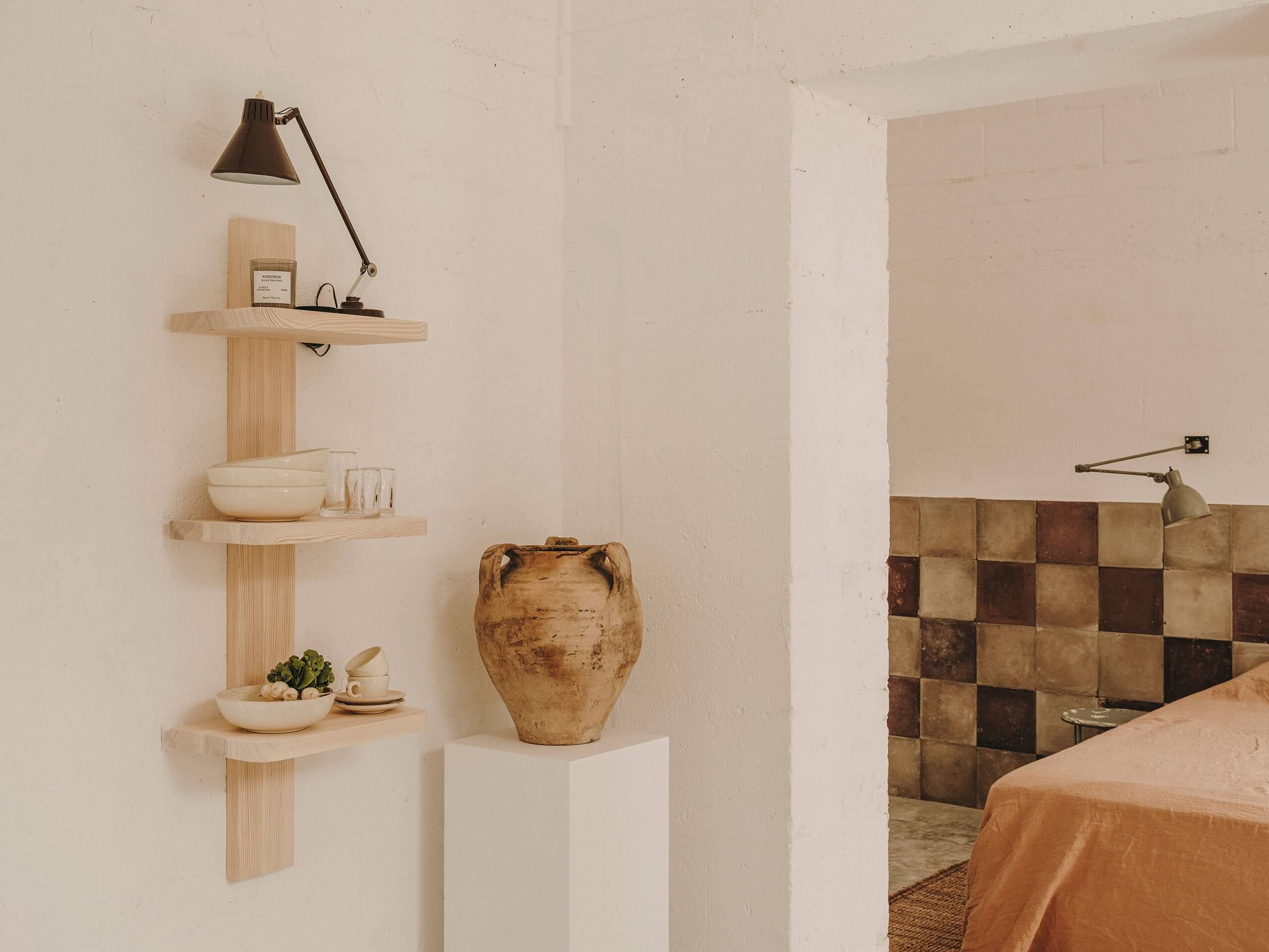 Borgo Gallana, Europe — Minimalist bedroom with natural wood floating shelves, terracotta pottery, checkered tile backsplash, and adjustable brass task lamp against whitewashed walls.
