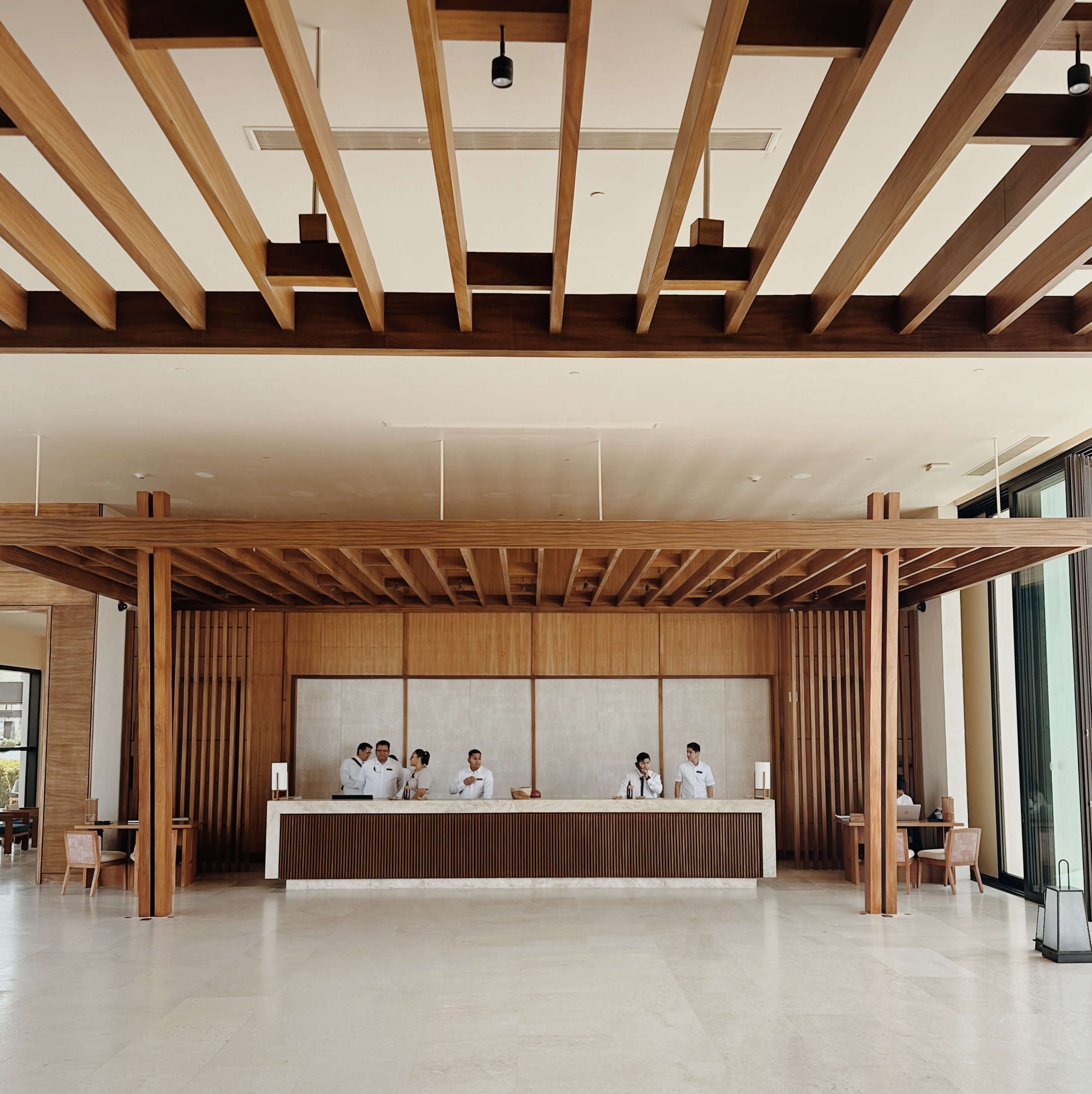 Nobu Los Cabos, Mexico — Minimalist hotel lobby with timber ceiling beams, polished concrete floor, and wooden reception desk with staff in white uniforms.
