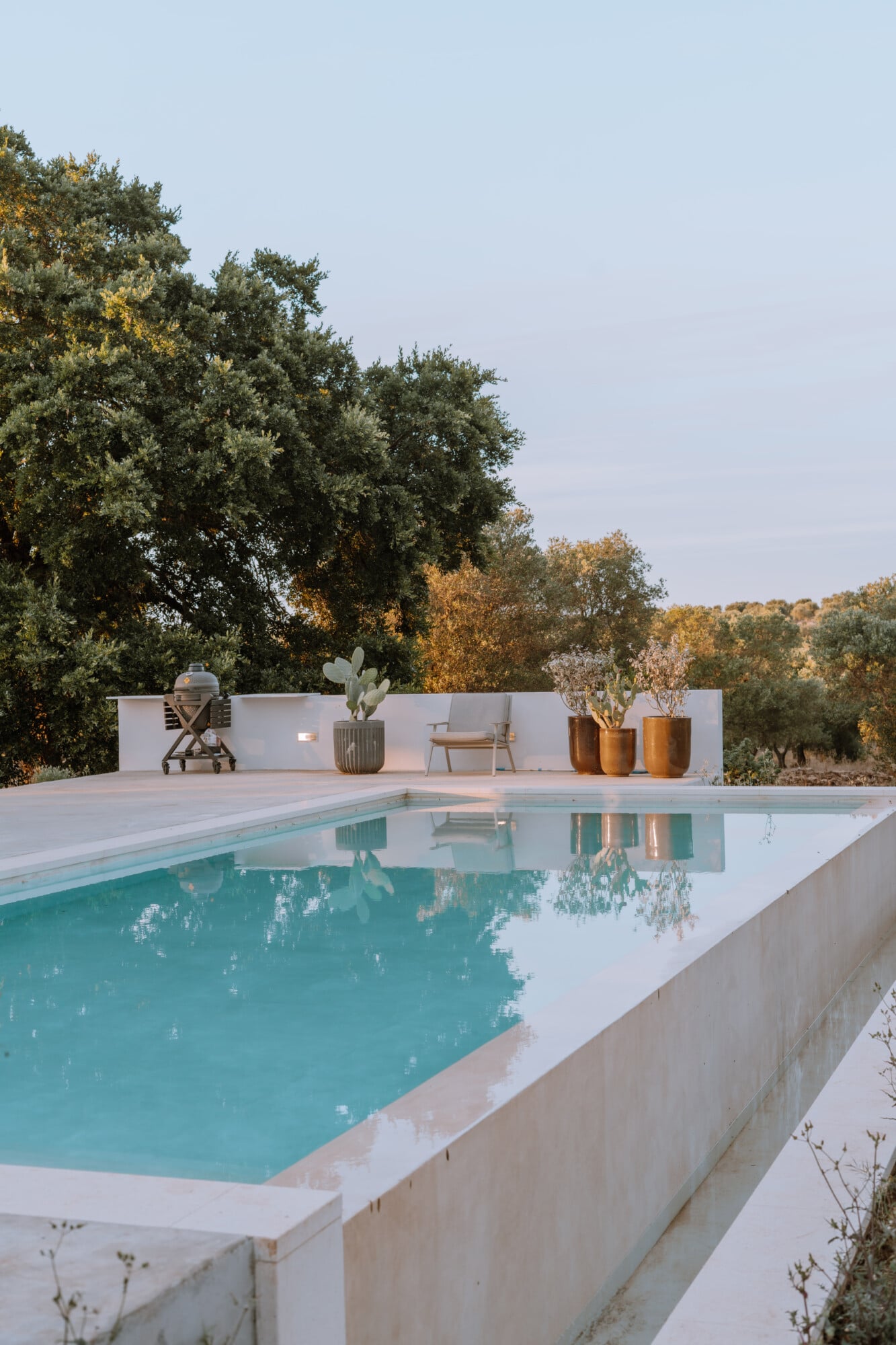 Villa Quercia, Italy — Modernist poolside with turquoise water, white stone terrace, minimalist white wall with potted cacti and wooden vessels, olive trees beyond in golden afternoon light.