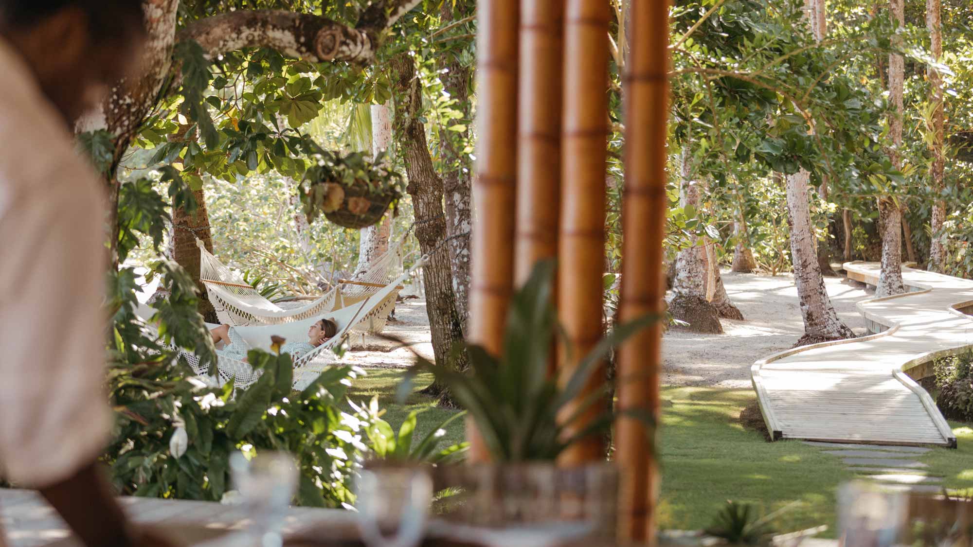 Islas Secas, Panama — Woman resting in cream hammock beneath palms and wooden pergola, dappled sunlight filtering through lush tropical foliage and boardwalk pathways.