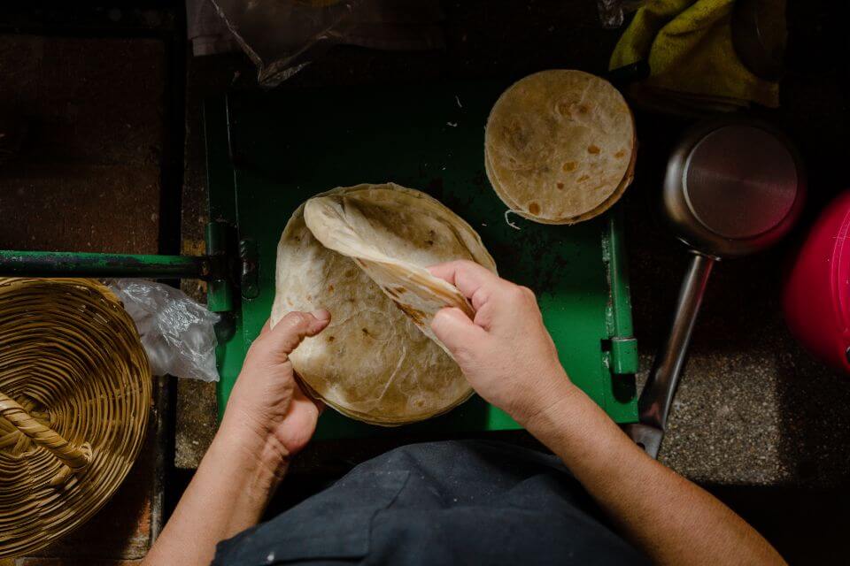 Hands fold a warm flour tortilla at Paradero Restaurant, surrounded by traditional Mexican dining essentials on a green surface.