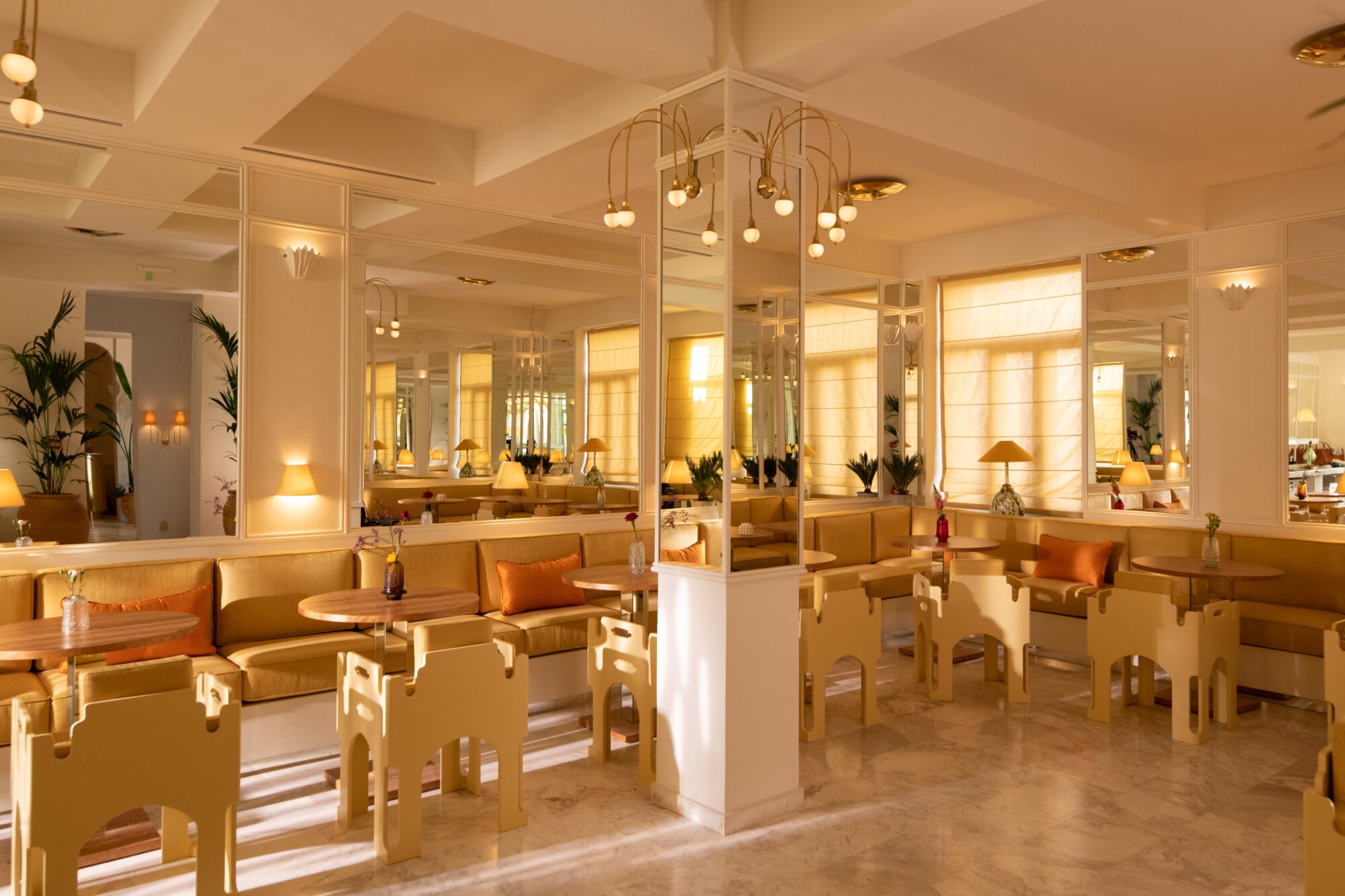 Apollo Palm, Europe — Sunlit dining room with natural wood furnishings, mirrored walls, brass pendant lights, and potted palms creating warm, elegant hospitality interior.