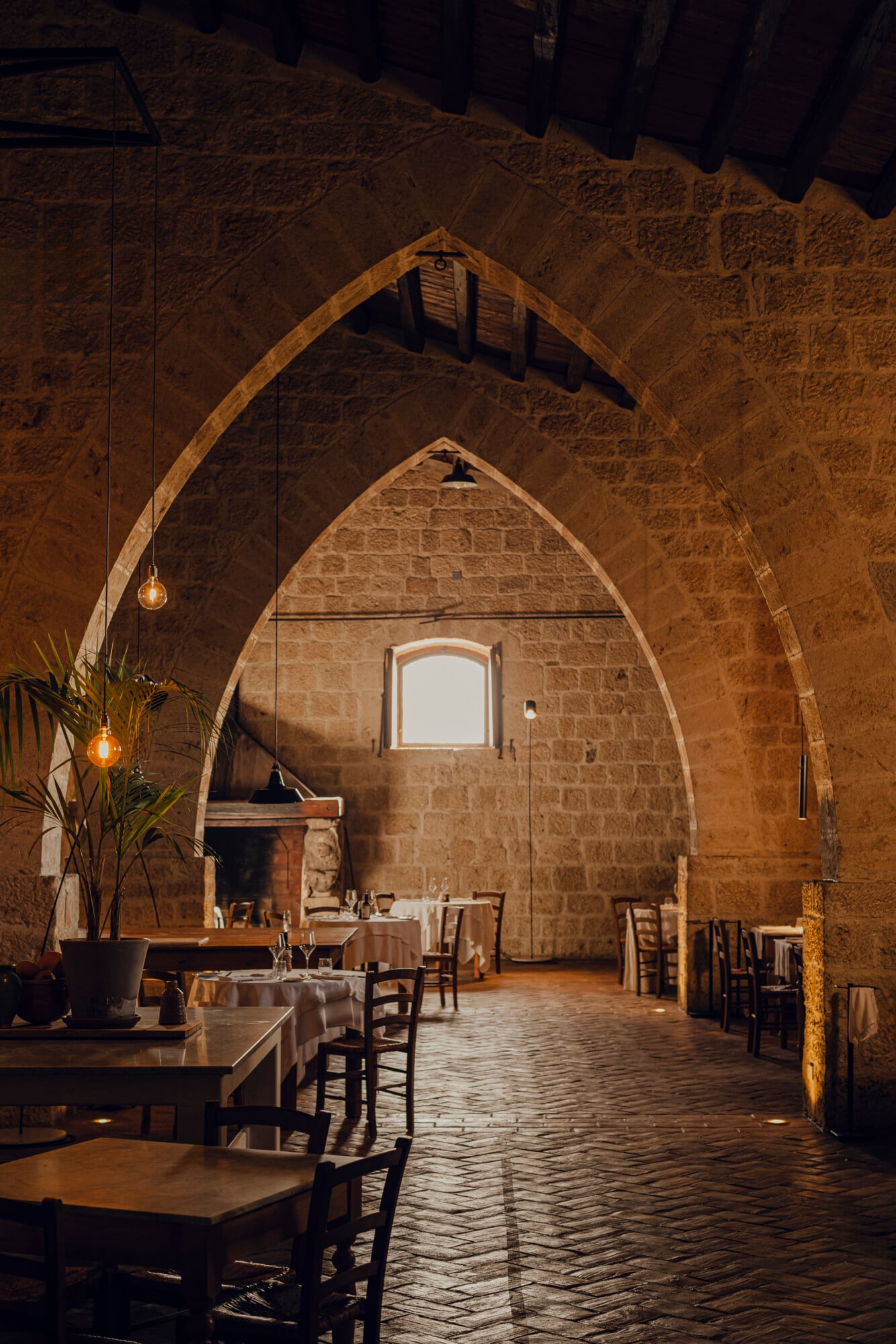 Susafa Restautant — Susafa Restaurant, Italy — Stone archway dining room with golden pendant lights, rustic wooden chairs, and natural window light casting warm shadows on brick walls.