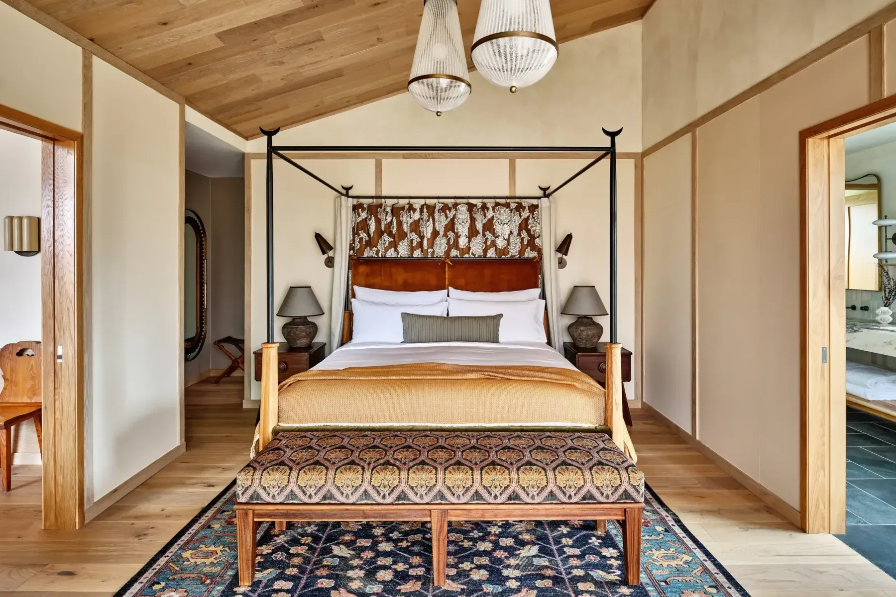 Wildflower Farms, North America — Canopy bed with black metal frame in light-filled bedroom with wood ceiling, cream walls, patterned headboard, and layered area rugs.