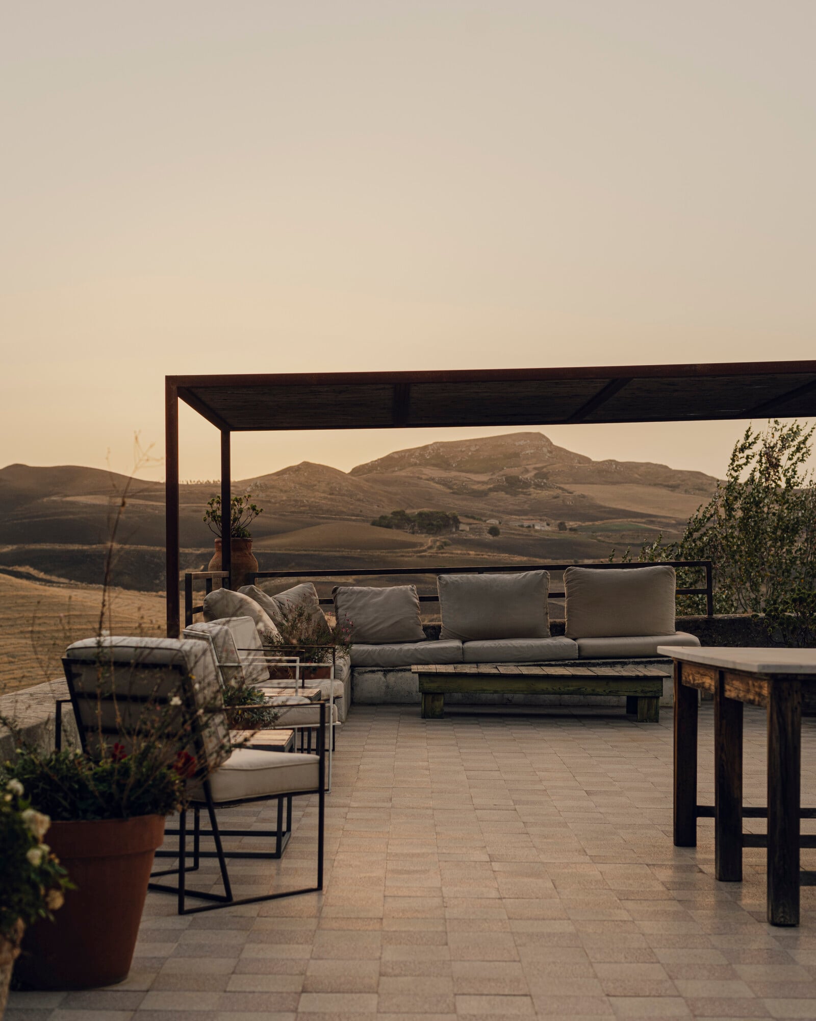 Susafa, Italy — Black pergola framing panoramic valley views, neutral linen seating, stone terrace at golden hour with distant mountains.