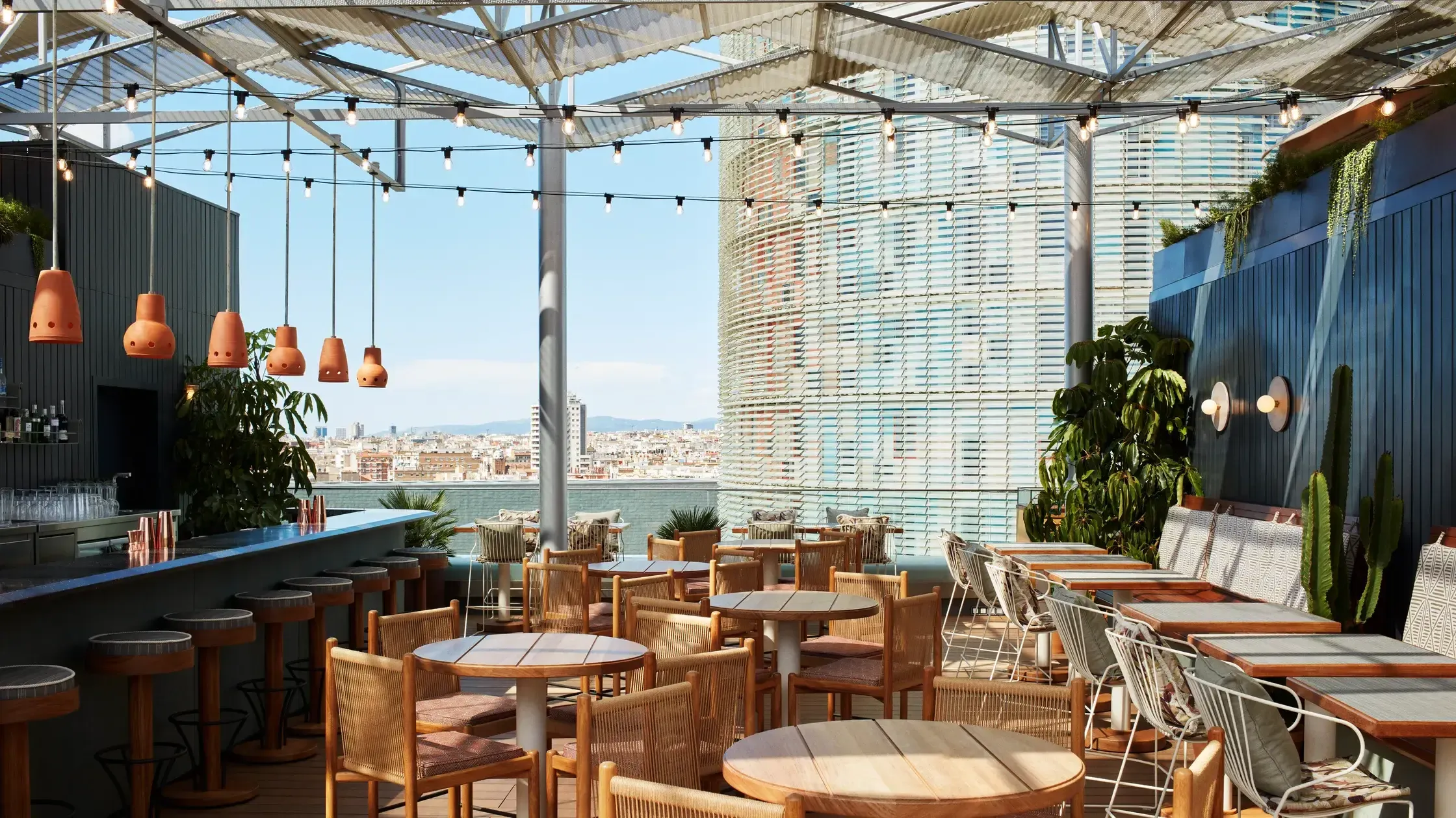 Tope, Europe — Rooftop bar terrace with woven wooden chairs, hanging copper pendants, shaded pergola, waterfront views, and potted greenery overlooking cityscape.