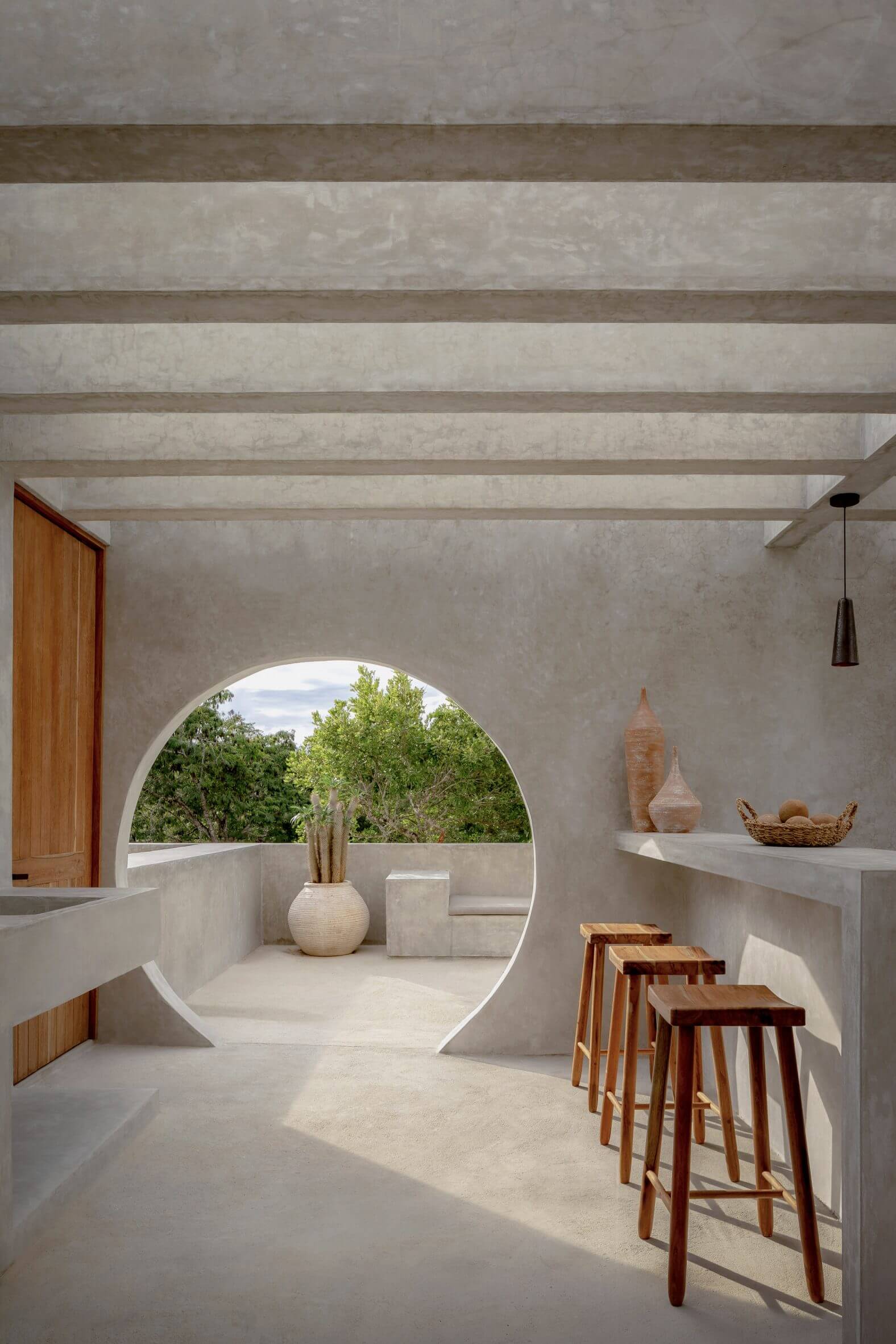 Villa Cava, Mexico — Minimalist concrete interior with circular archway framing lush landscape, wooden bar stools, and terracotta vessels on shelving.