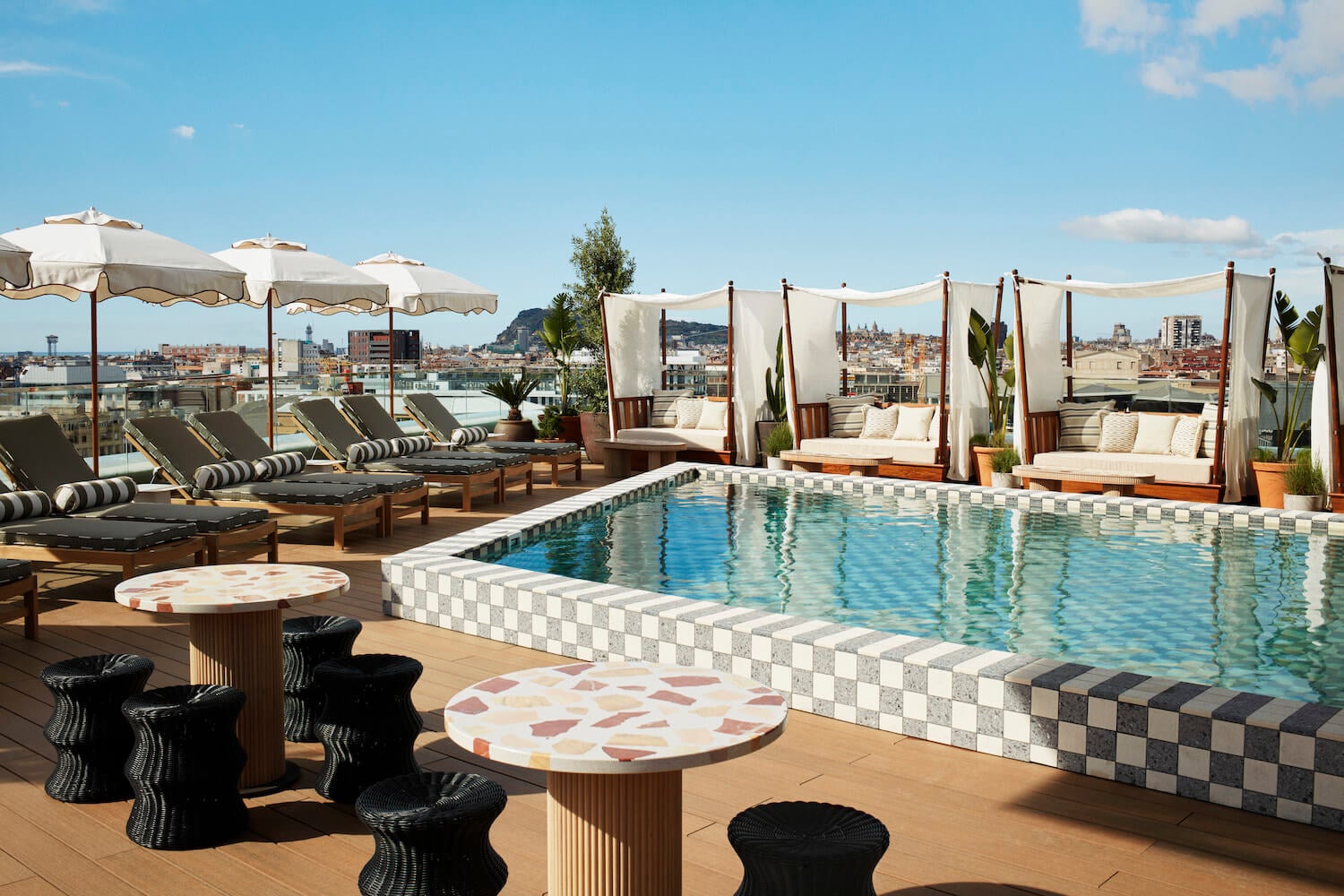 The Hoxton Poblenou, Barcelona, Spain — rooftop pool with checkered tile border, striped loungers, canopied daybeds, and city skyline views under clear sky.