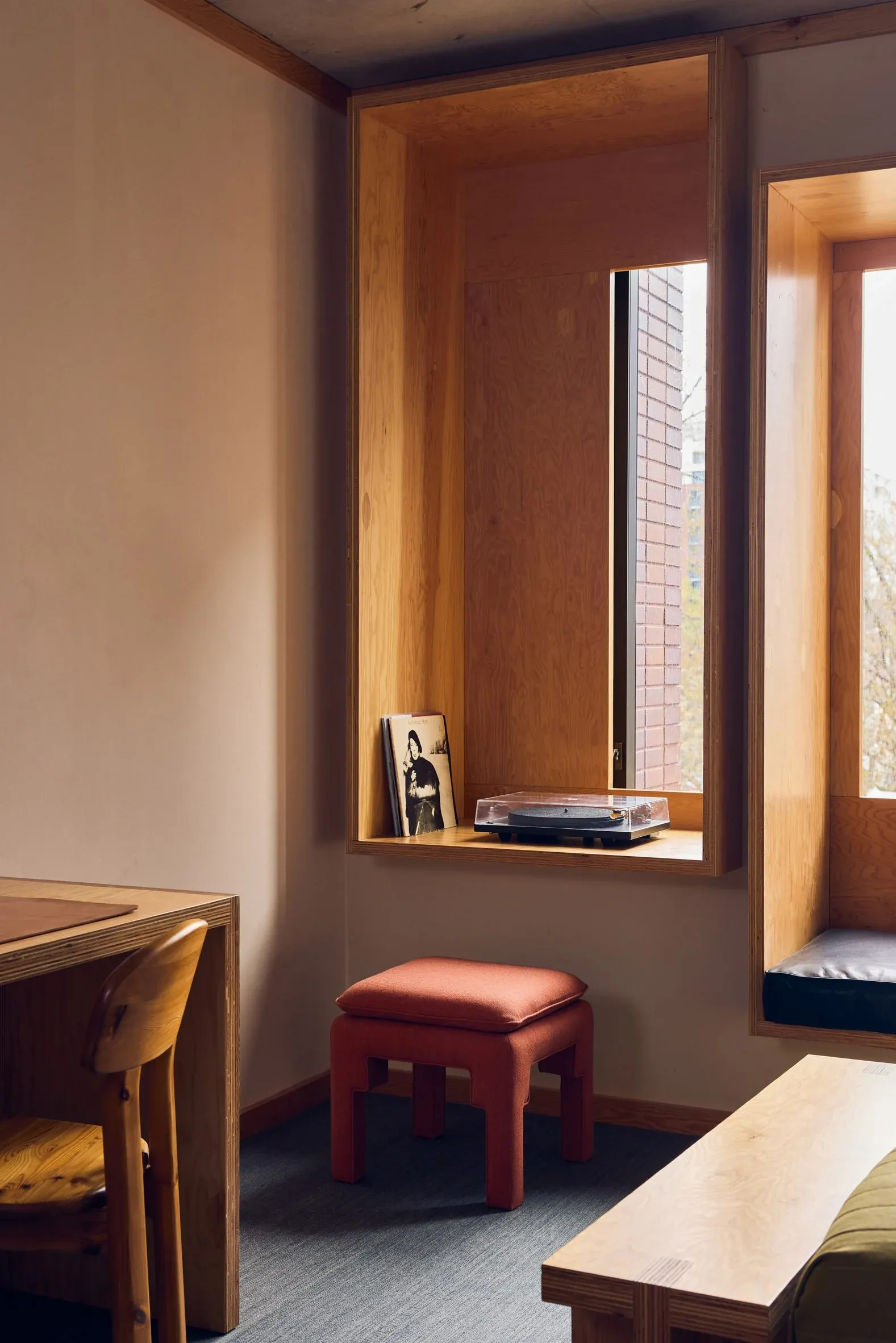 Ace Hotel Toronto, Canada — Warm timber-framed window nook with coral upholstered stool, natural light, minimalist shelving, and brick exterior view.