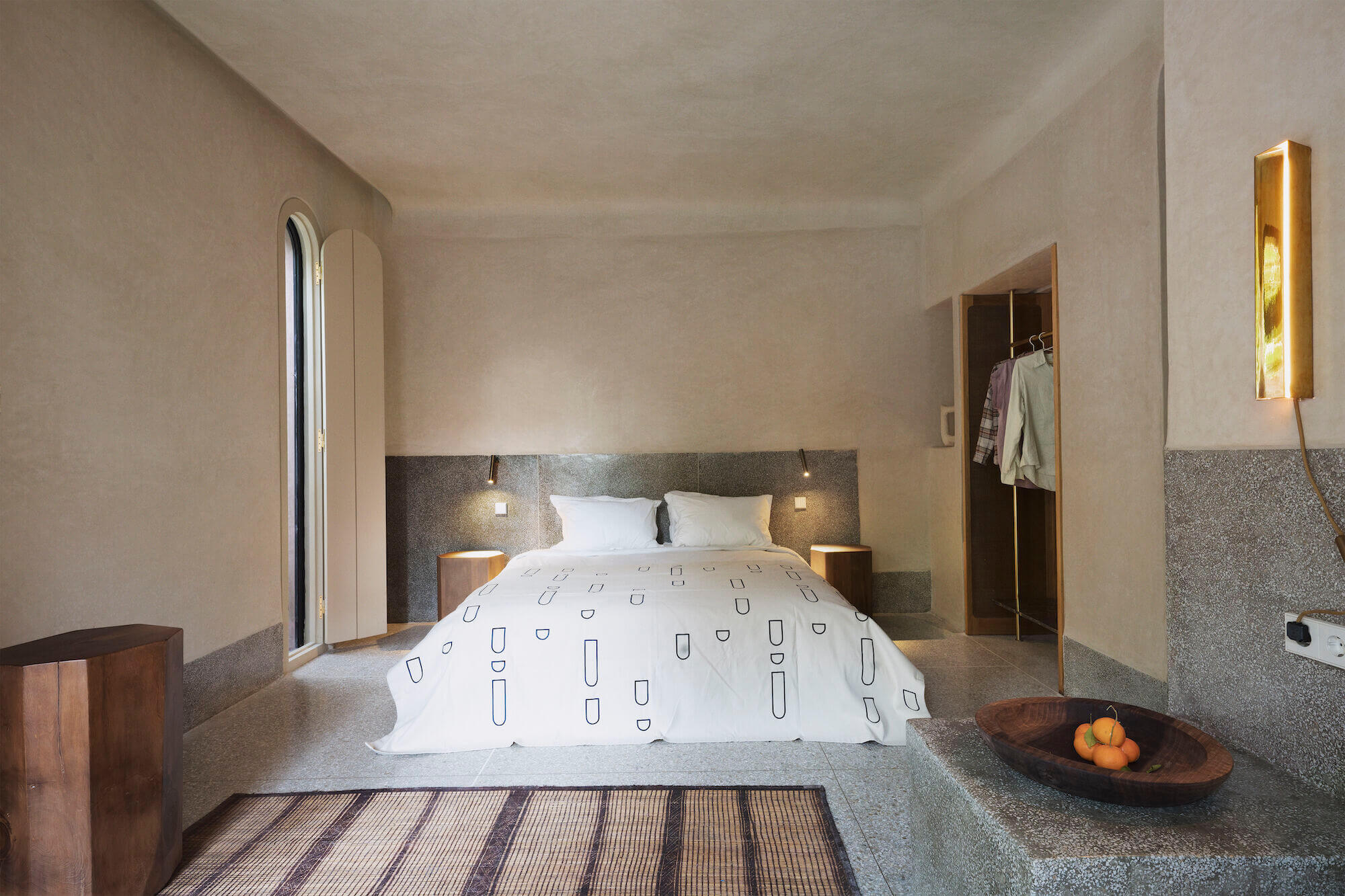 Maison Brummell Majorelle, Africa — Minimalist bedroom with terrazzo flooring, dark stone headwall, brass bedside sconces, and natural light from arched window.