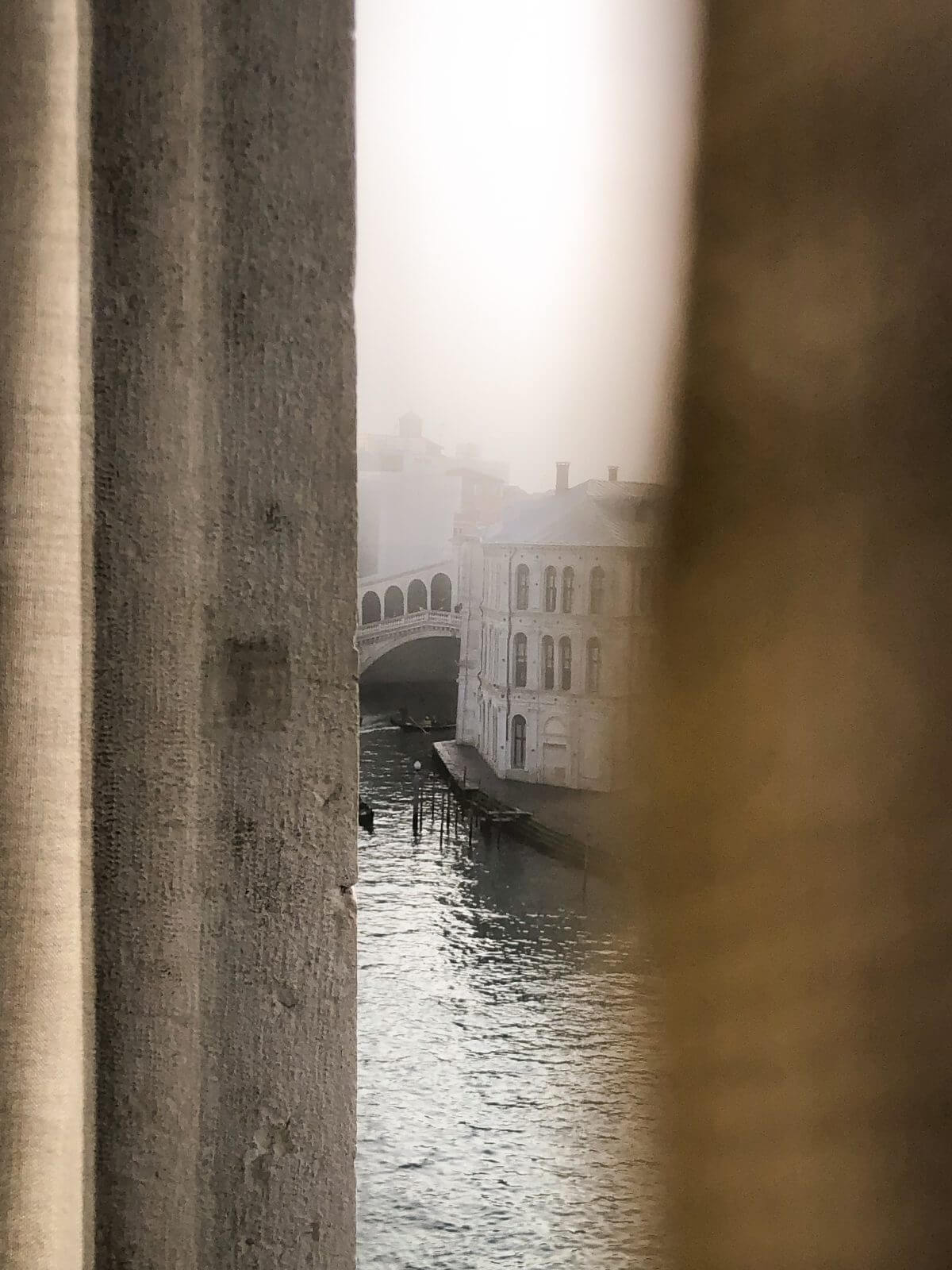 The Venice Venice Hotel, Venice, Italy — Venetian canal framed through stone archway with Renaissance bridge and palazzo facade emerging from mist