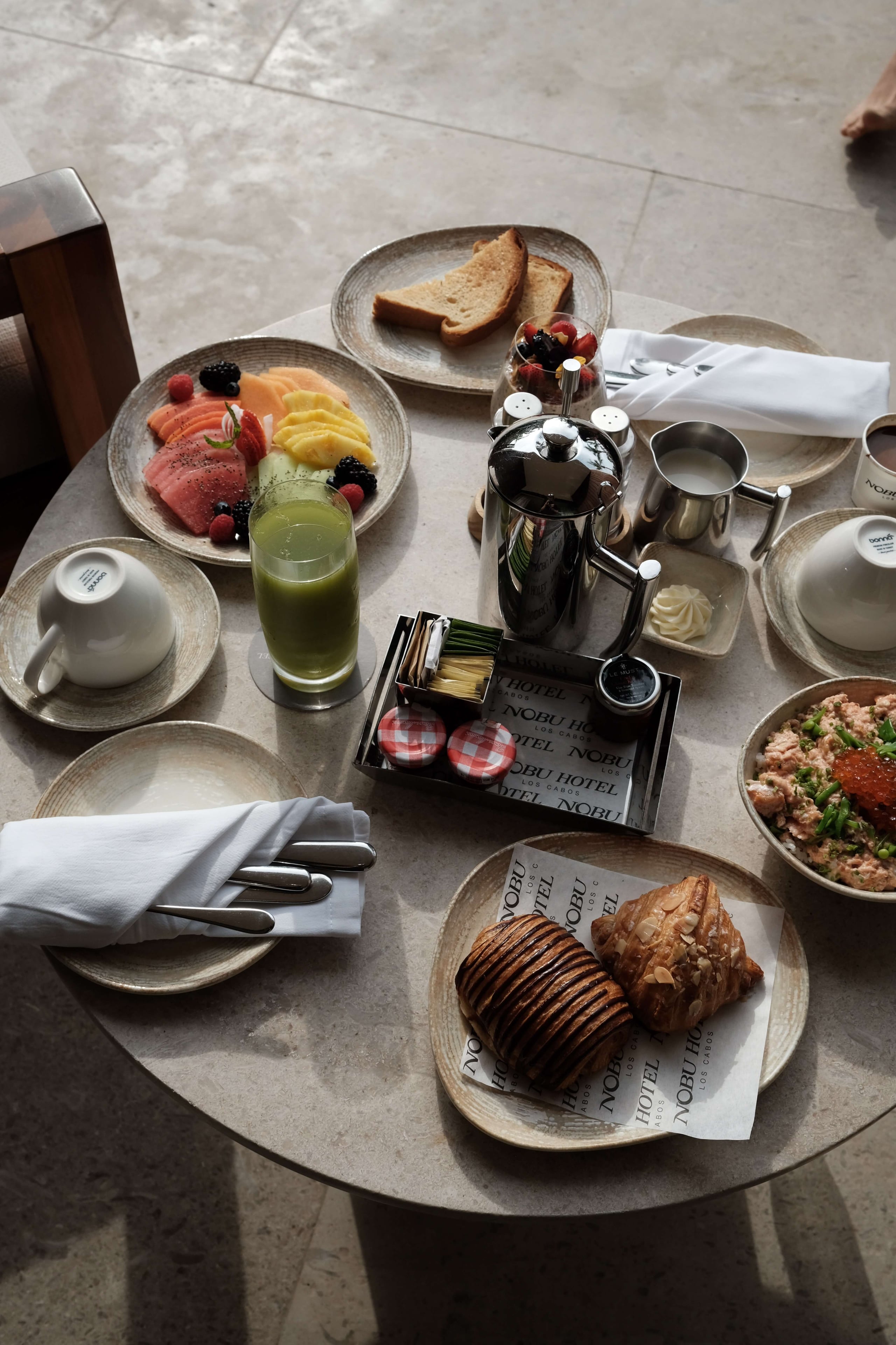 Nobu Los Cabos, Mexico — Overhead view of luxury breakfast table with sashimi platter, pastries, fresh juice, and minimalist tableware on stone surface.
