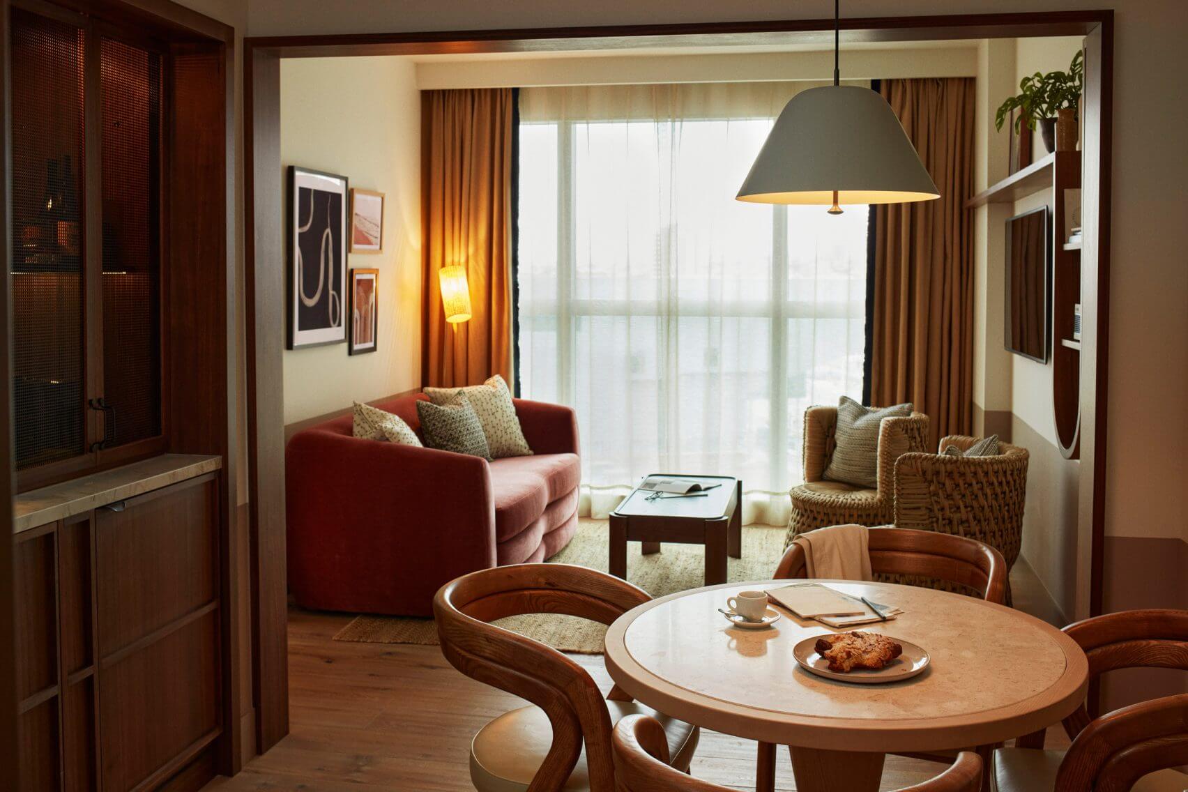 The Hoxton Poblenou, Barcelona, Spain — Spacious hotel suite with wooden dining table, red curved sofa, woven armchairs, pendant lamp, and floor-to-ceiling windows overlooking city.
