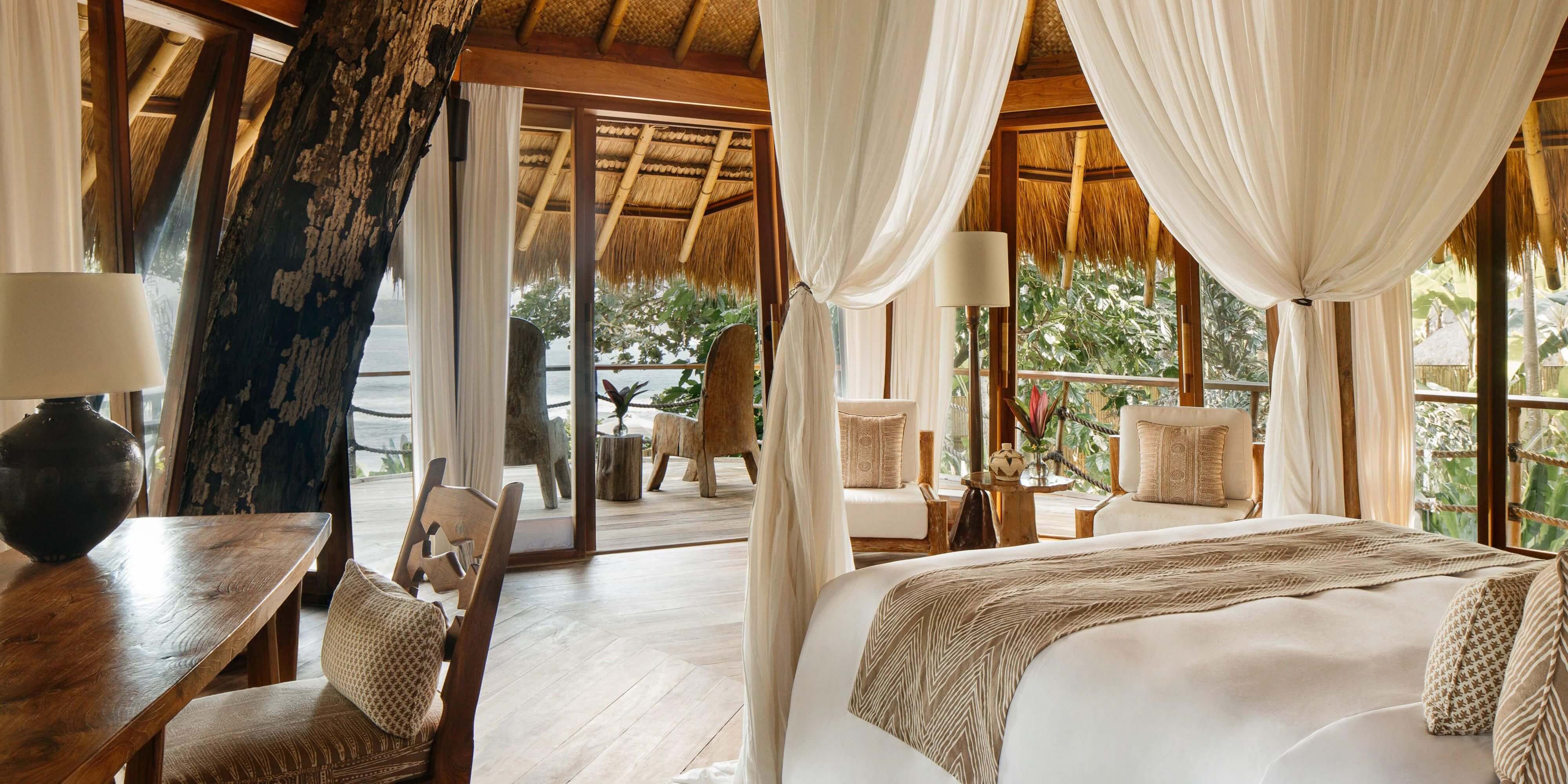 NIHI Sumba, Indonesia — Oceanfront villa bedroom with draped canopy bed, thatched roof, wooden furnishings, and open terrace overlooking tropical garden and sea.