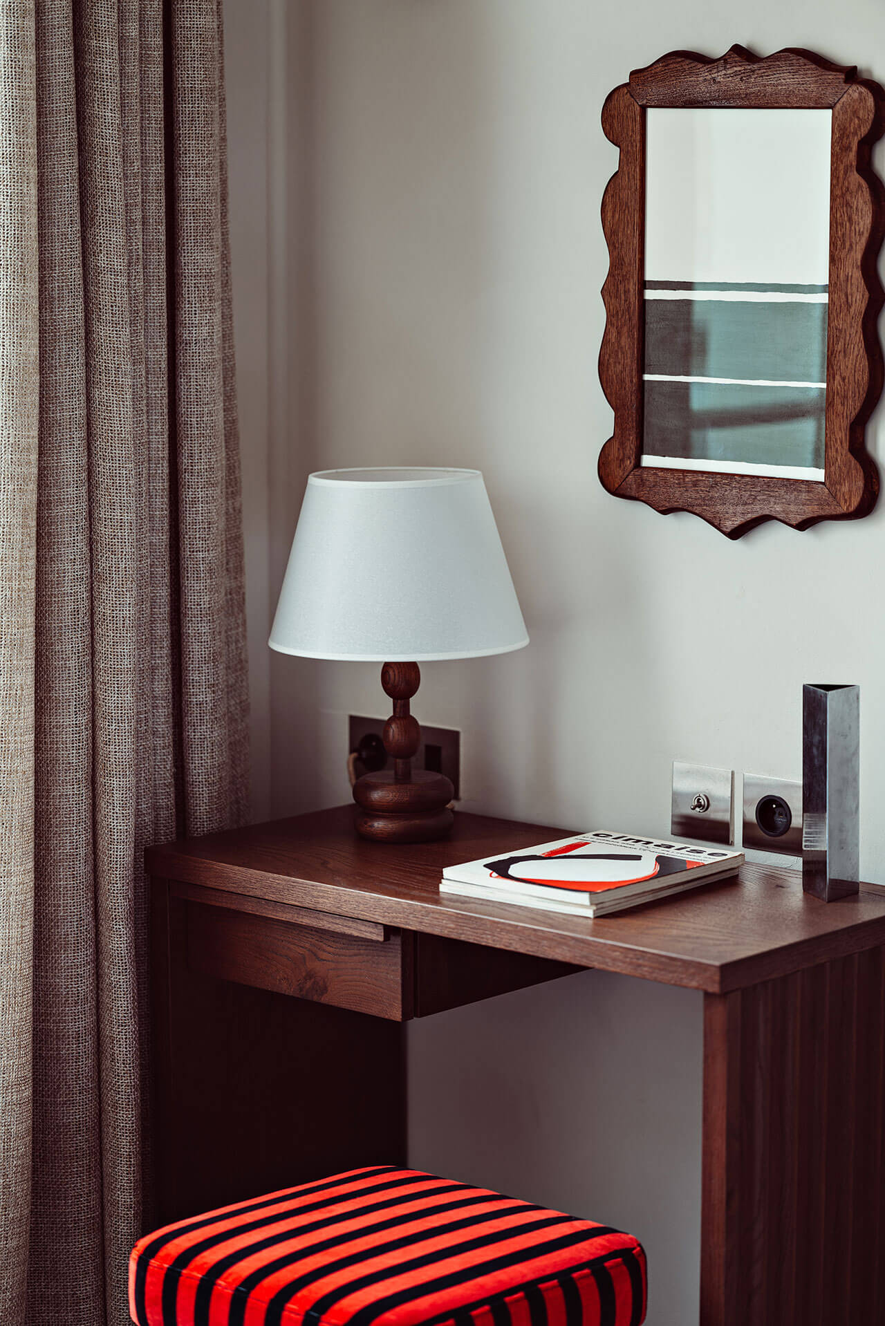 Académies et des Arts, Europe — Wooden desk with white table lamp, decorative mirror, striped ottoman, and textured curtains in warm, sophisticated interior.