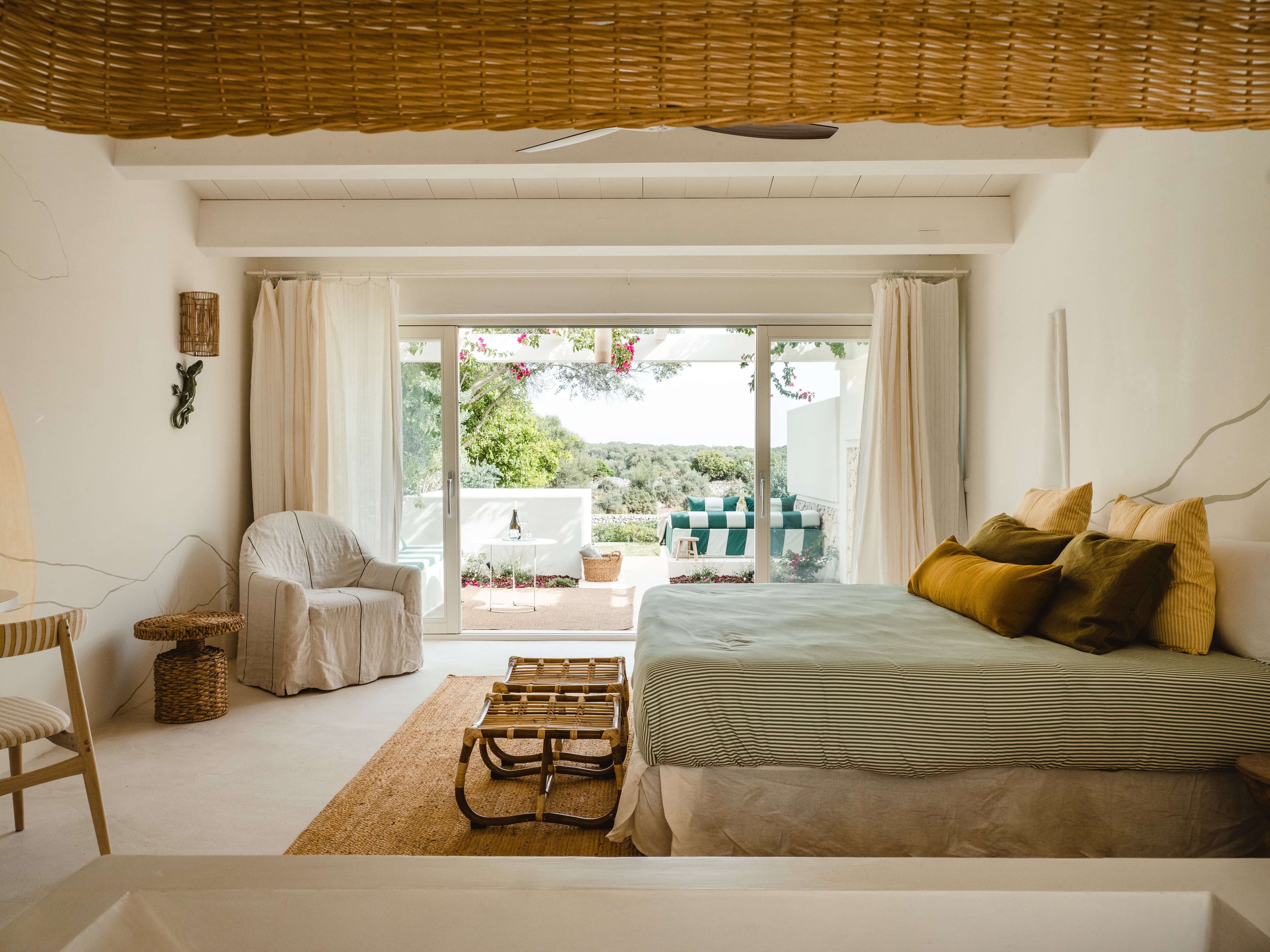 Morvedra Nou, Balearic Islands — Bright bedroom with striped bedding, woven ceiling, cream linen chair, and expansive glass doors opening to Mediterranean landscape views.