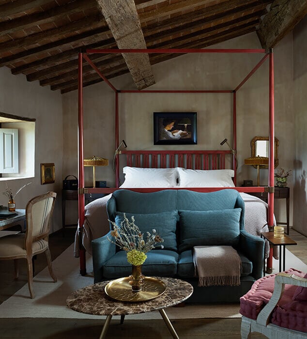 Castello di Reschio, Europe — Four-poster bed with red frame in rustic bedroom with exposed wooden beams, teal velvet sofa, marble side table, and period furnishings.