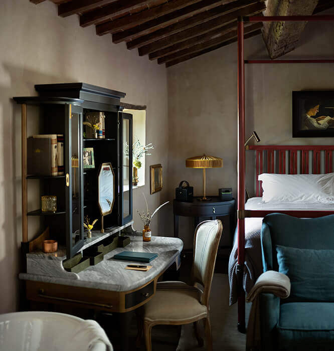 Castello di Reschio, Europe — Luxe bedroom suite with marble vanity, black display cabinet, red four-poster bed, exposed wooden beams, and muted taupe walls.