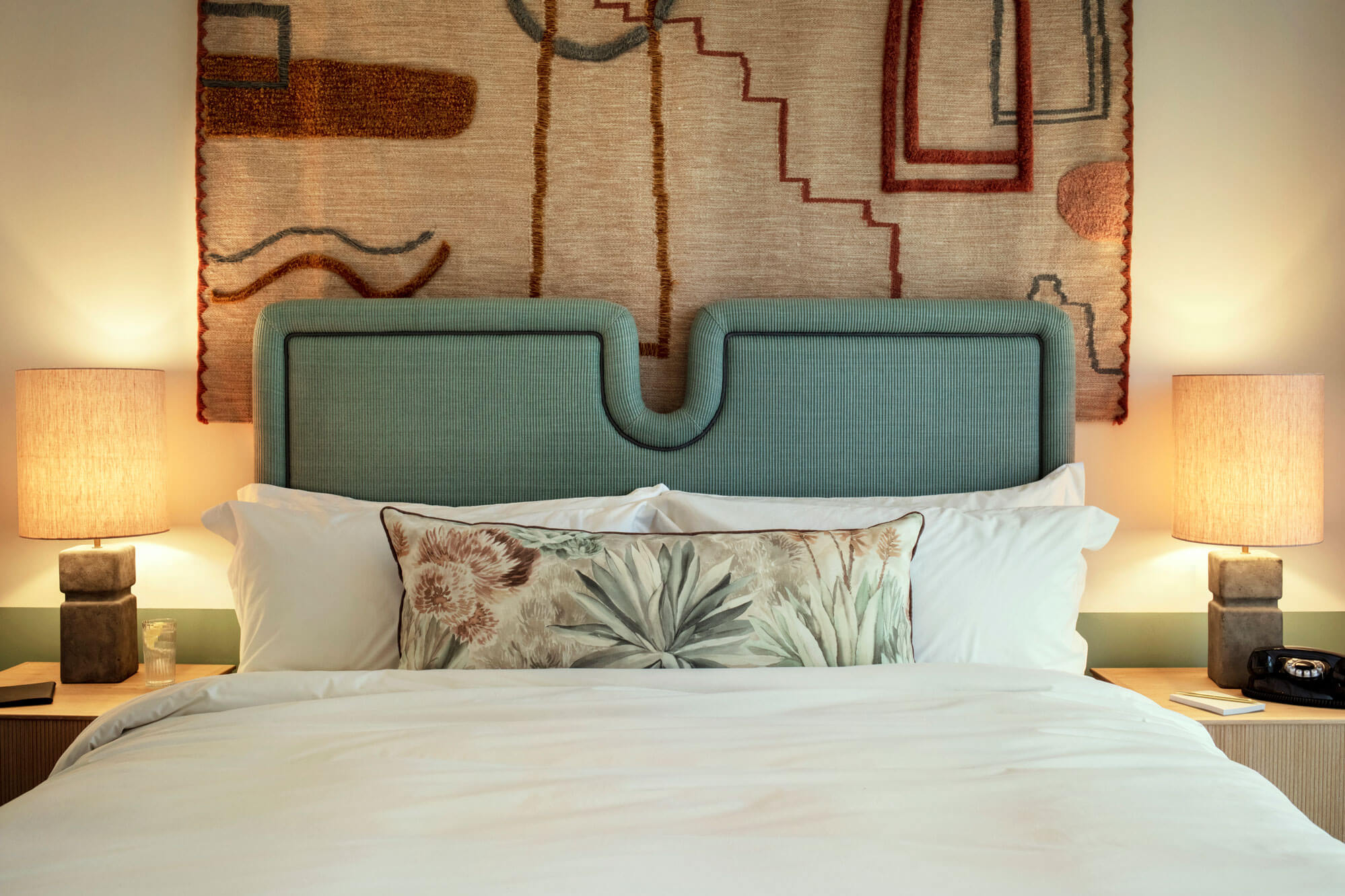 The Hoxton Poblenou, Barcelona, Spain — Serene hotel bedroom with seafoam upholstered headboard, white linens, botanical pillow, and abstract tapestry above warm brass bedside lamps.