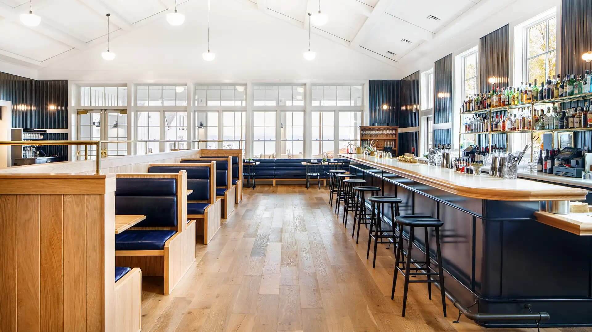 The Lake House, North America — Modern lakeside bar with vaulted white ceiling, natural wood booths, navy leather seating, and expansive gridded windows overlooking water.