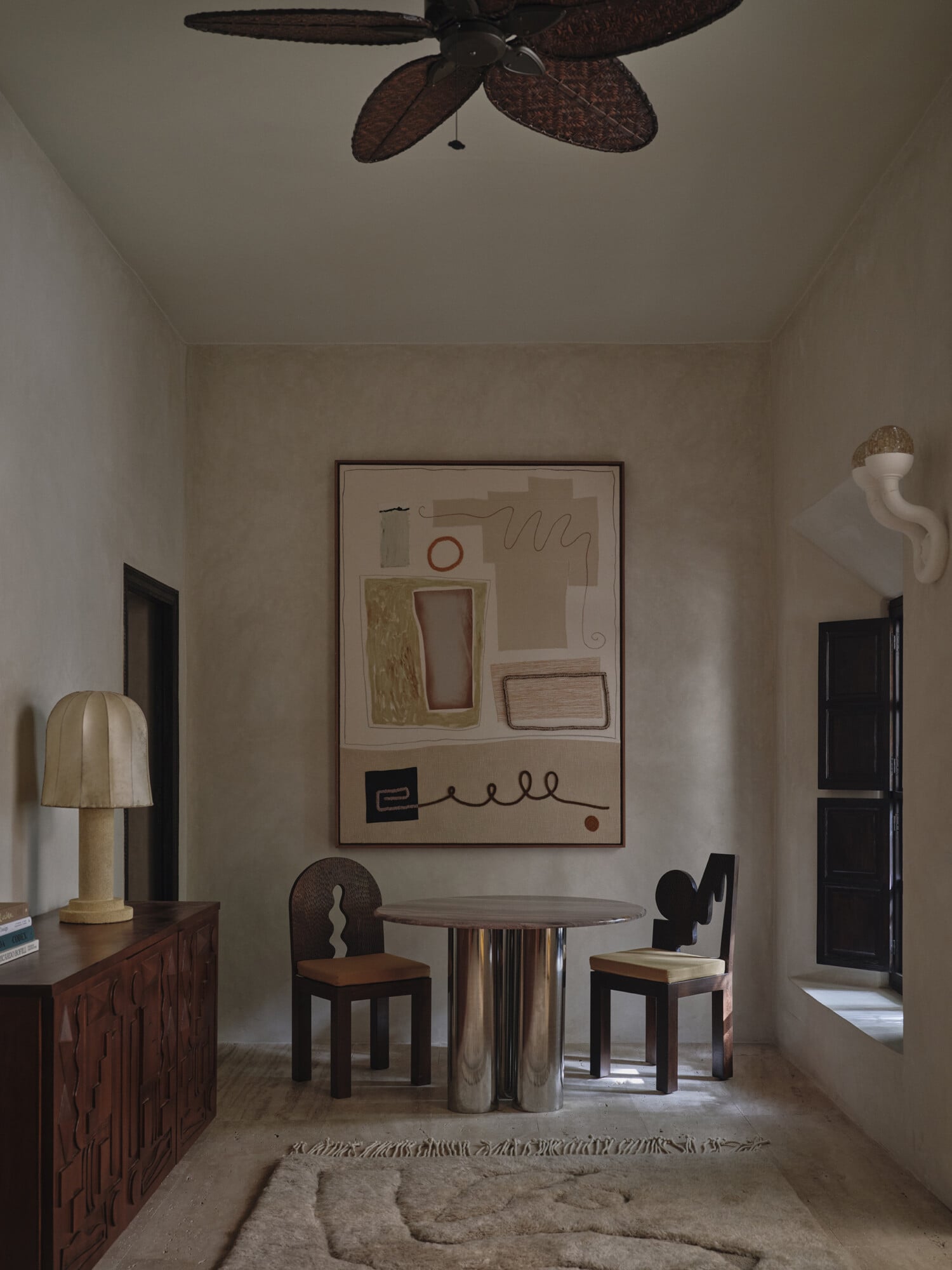 Rosemary, Africa — Minimalist interior with cream walls, dark wood furniture, geometric artwork, and sculptural decor beneath woven ceiling fan.