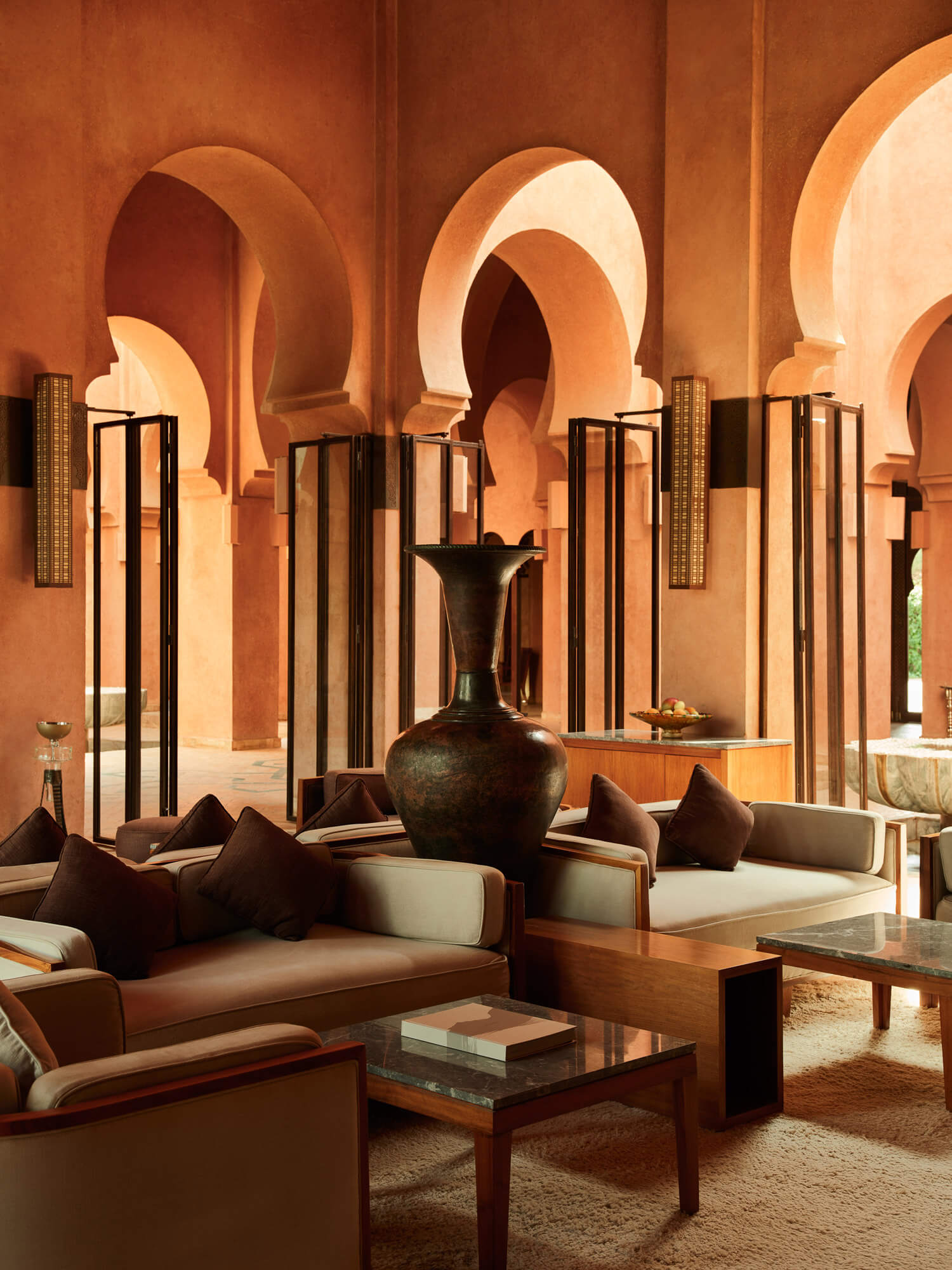 Amanjena, Marrakech, Morocco — Terracotta arches frame lounge seating with dark cushions and carved wooden details beneath sculptural lighting.