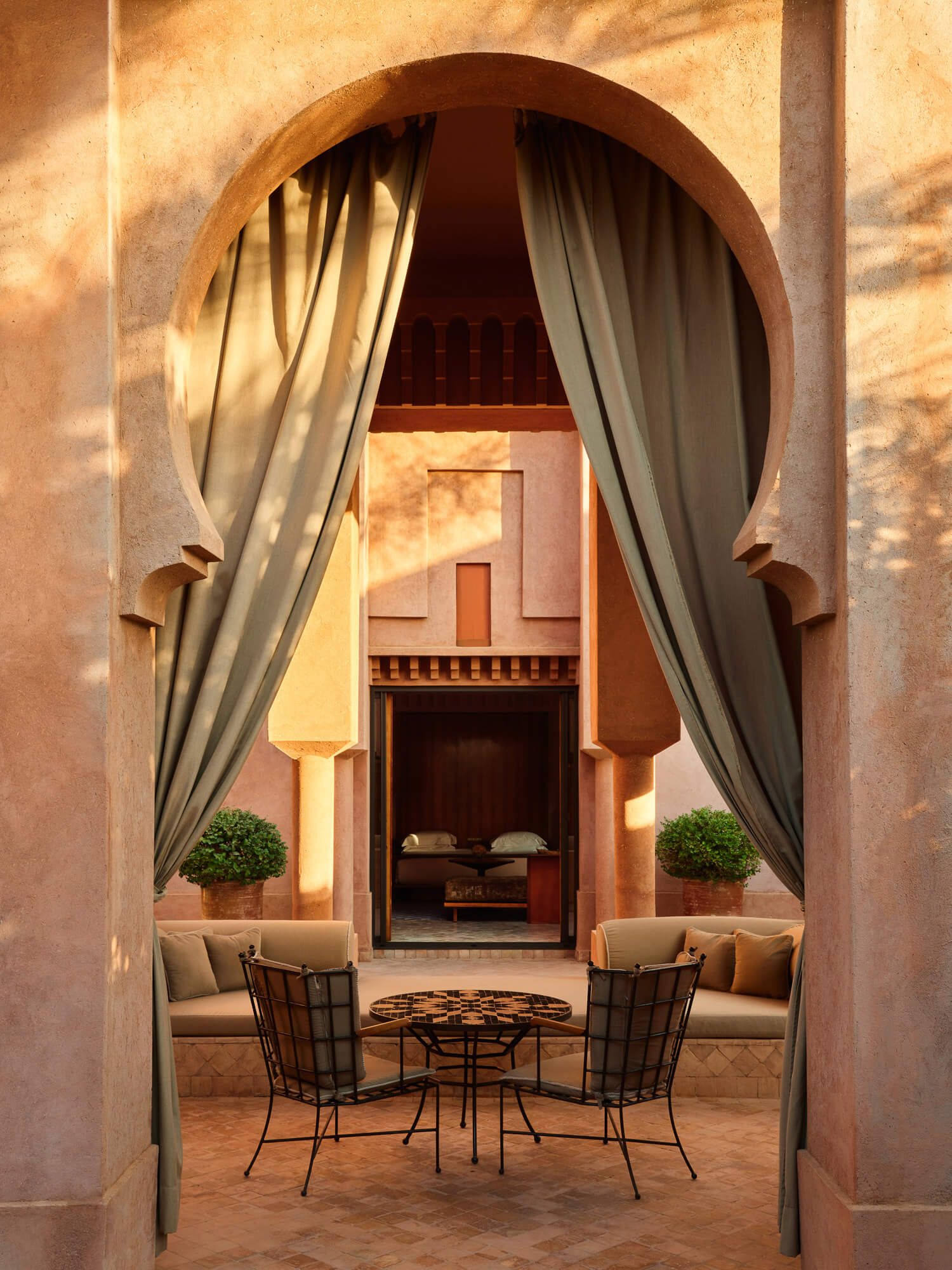 Amanjena, Africa — Arched courtyard with sheer curtains, wrought-iron seating, zellige table, warm terracotta walls and Moroccan architectural detail.