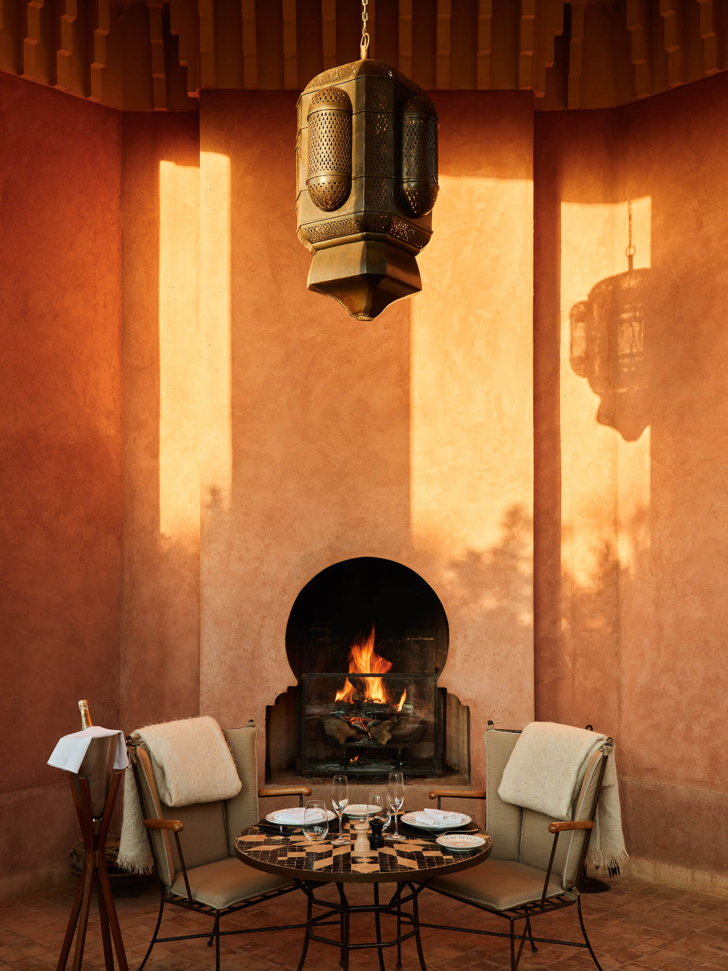 Amanjena, Africa — Ochre courtyard with arched fireplace, perforated brass lantern, geometric tile table, and linen chairs in warm evening light.