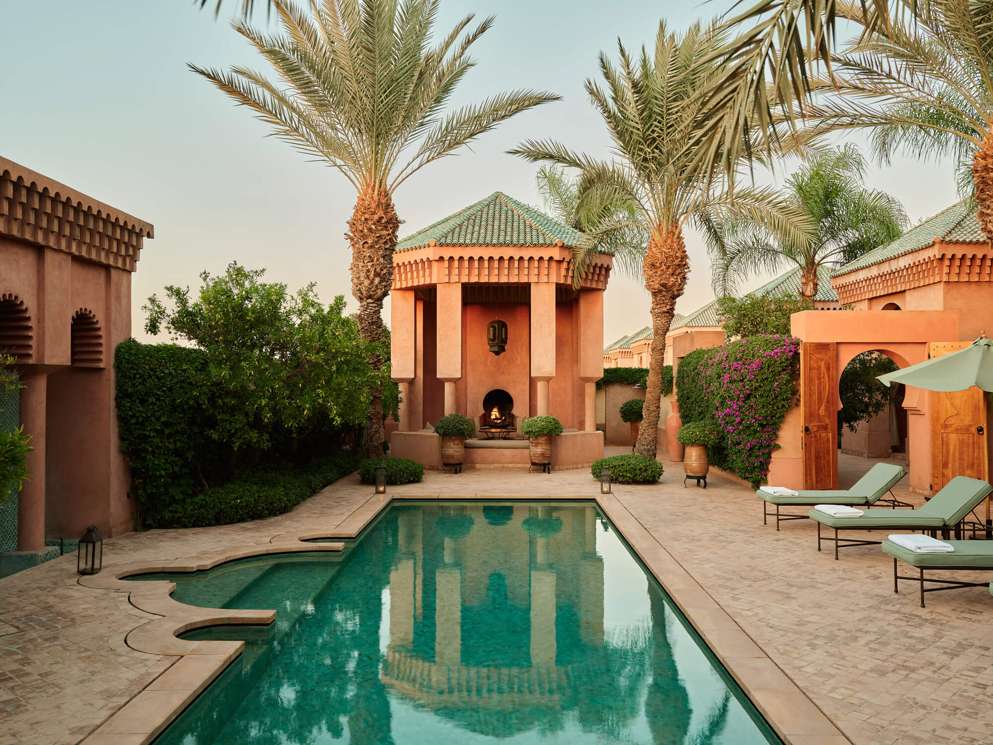 Amanjena, Marrakech, Morocco — Terracotta riad with central pool reflecting geometric architecture, palm trees, and magenta bougainvillea in desert courtyard at dusk.