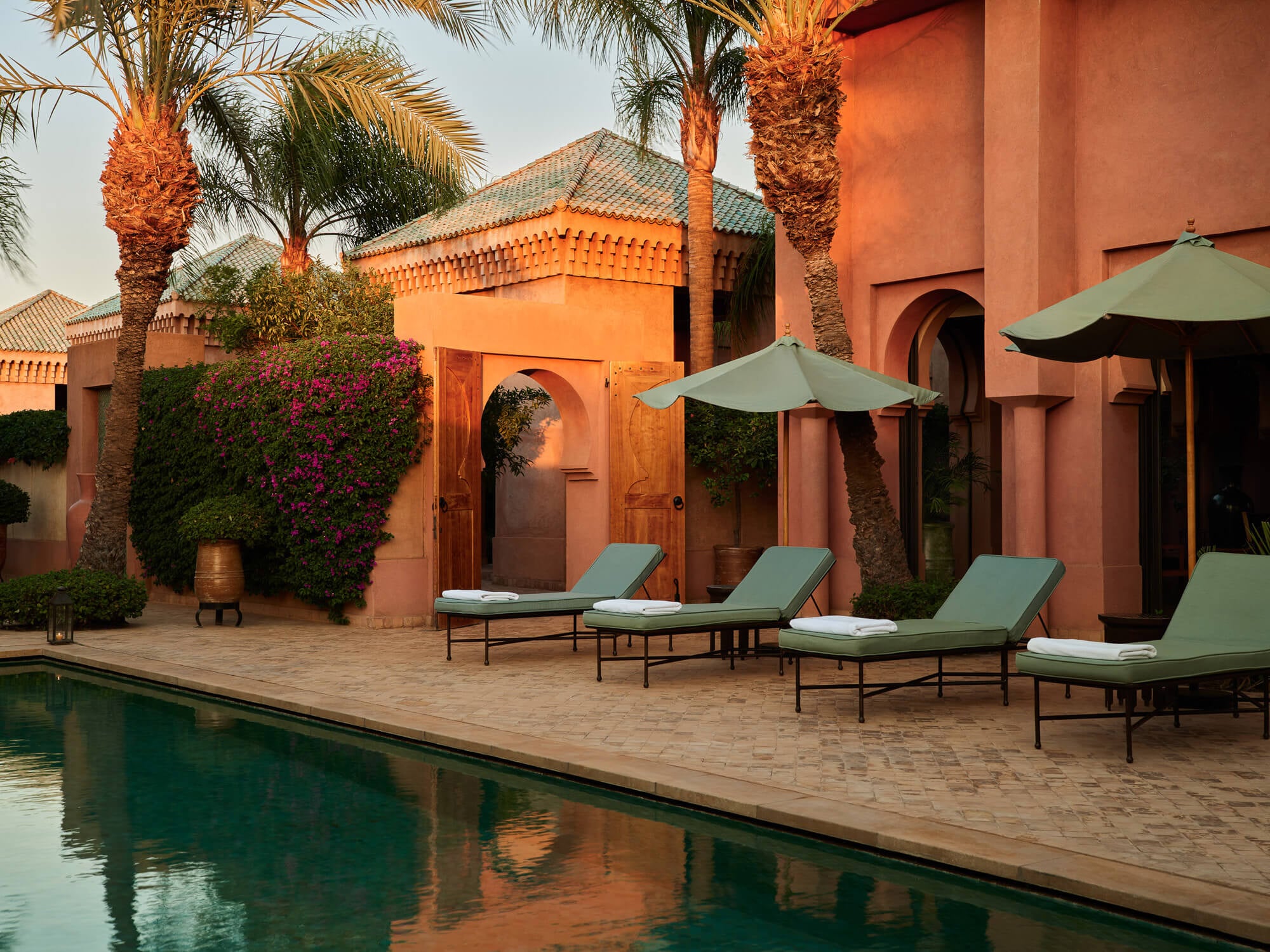 Amanjena, Africa — Terracotta riad with green sun loungers beside still pool, palm trees and magenta bougainvillea in afternoon light.